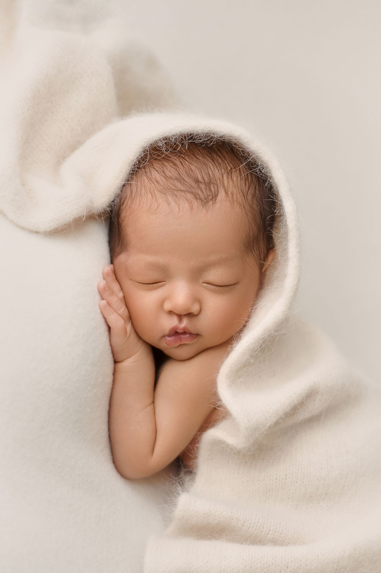 Peaceful newborn baby sleeping wrapped in a soft white knit blanket with hands tucked under cheek.