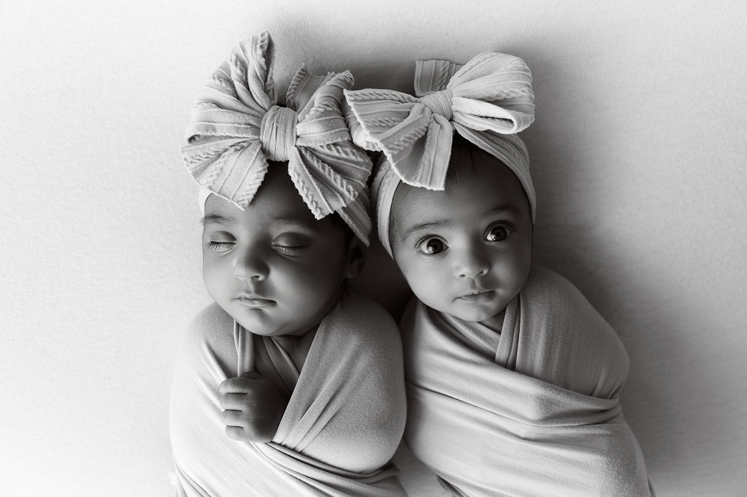 Two newborn twins swaddled in white wraps wearing striped bow headbands pose together in a black and white photo.