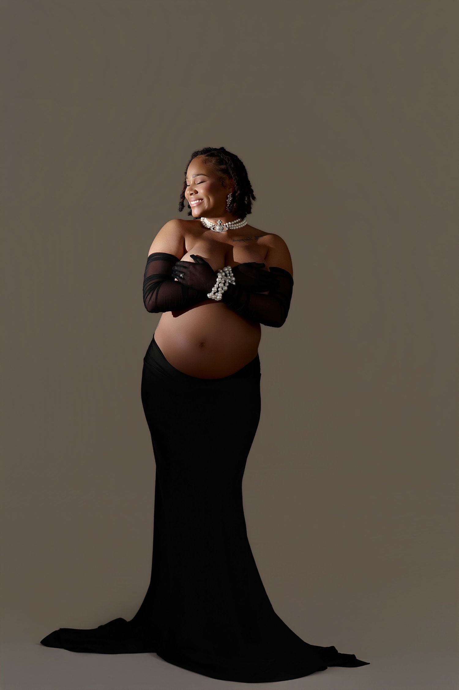 Pregnant woman in black off-shoulder gown with sheer gloves, cradling baby bump, smiling, studio maternity portrait.