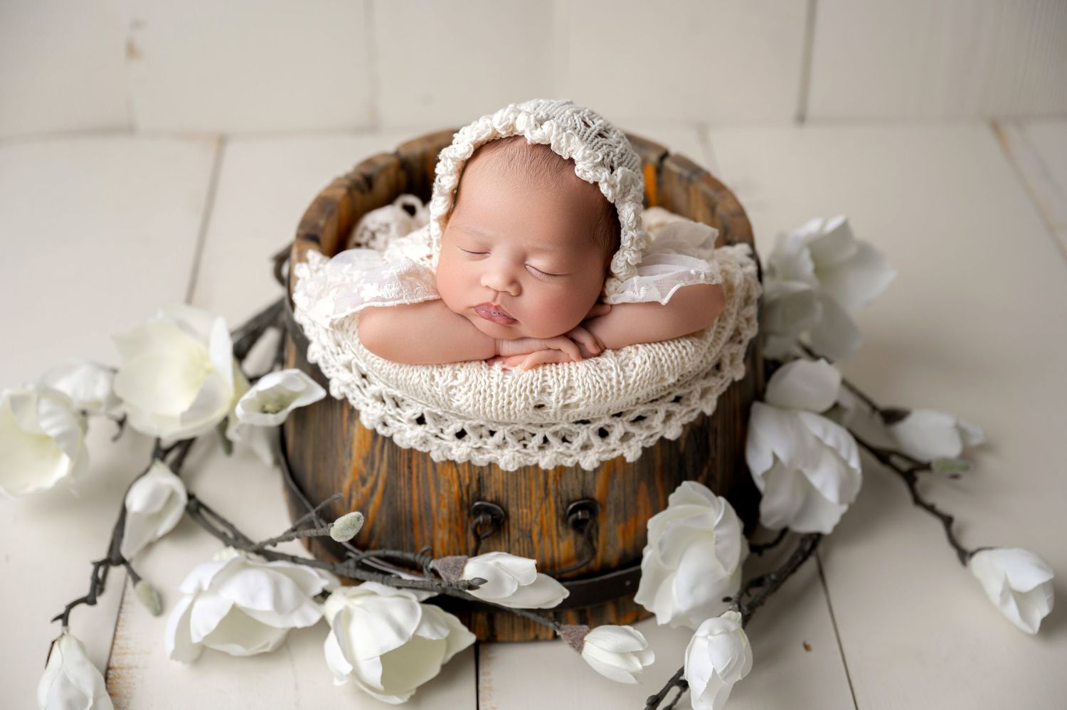 Sleeping newborn baby in rustic wooden bucket with lace bonnet, surrounded by white flowers on white wood floor.