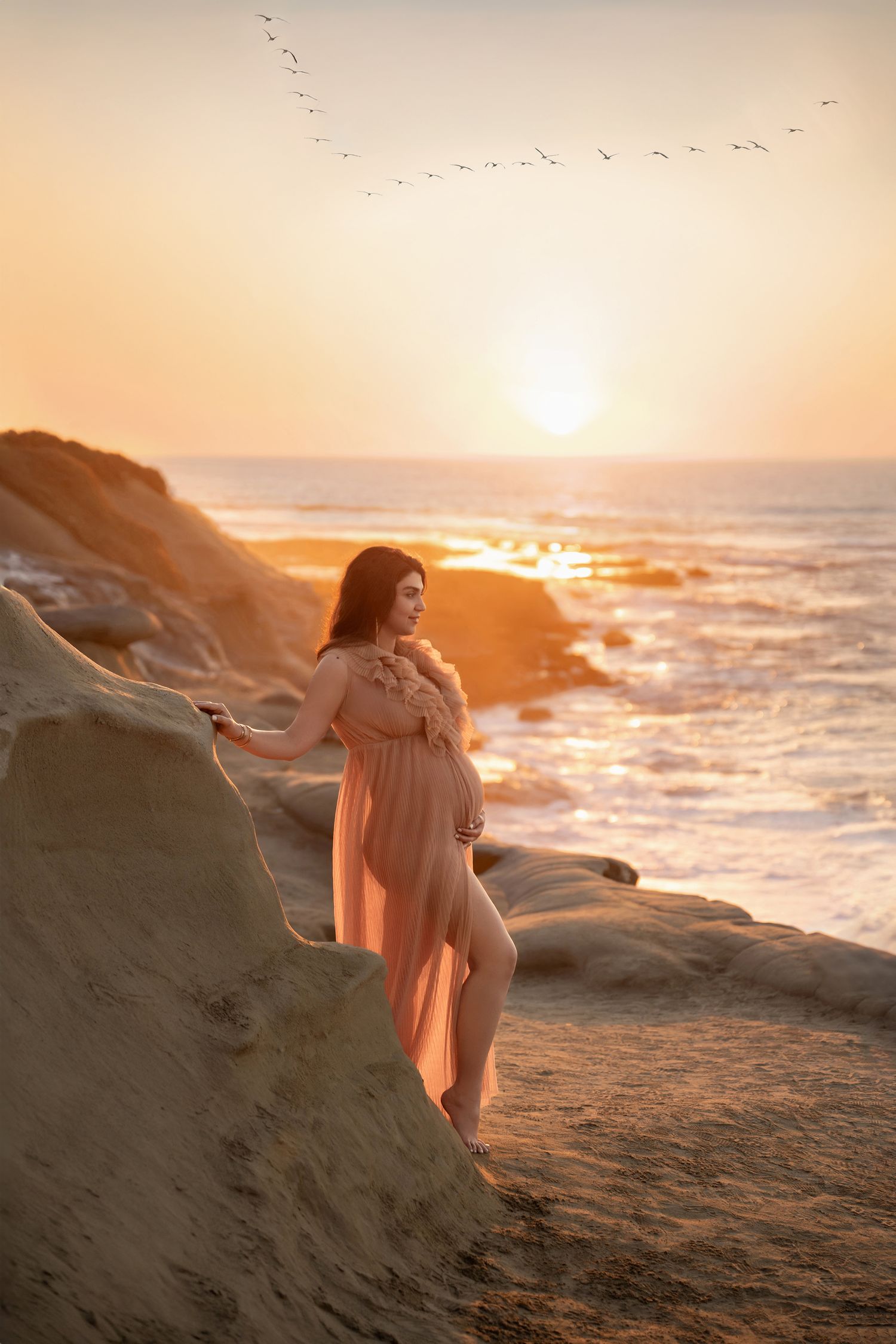 Pregnant woman in flowing peach gown poses on coastal rocks during golden sunset with birds flying overhead.