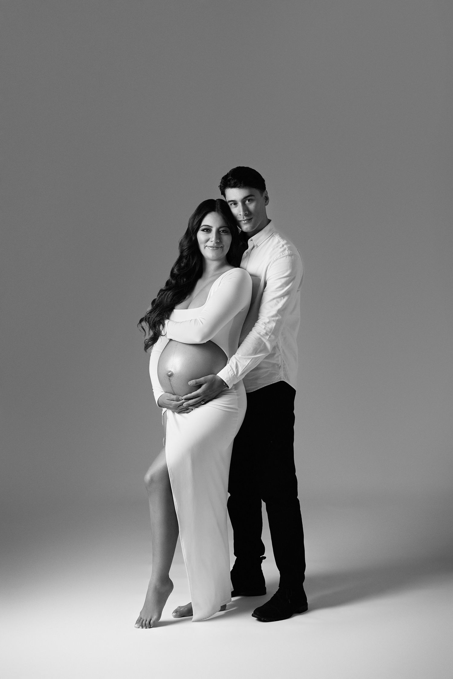 Black and white maternity photo of a couple, man embracing pregnant woman in white dress, both smiling at camera.