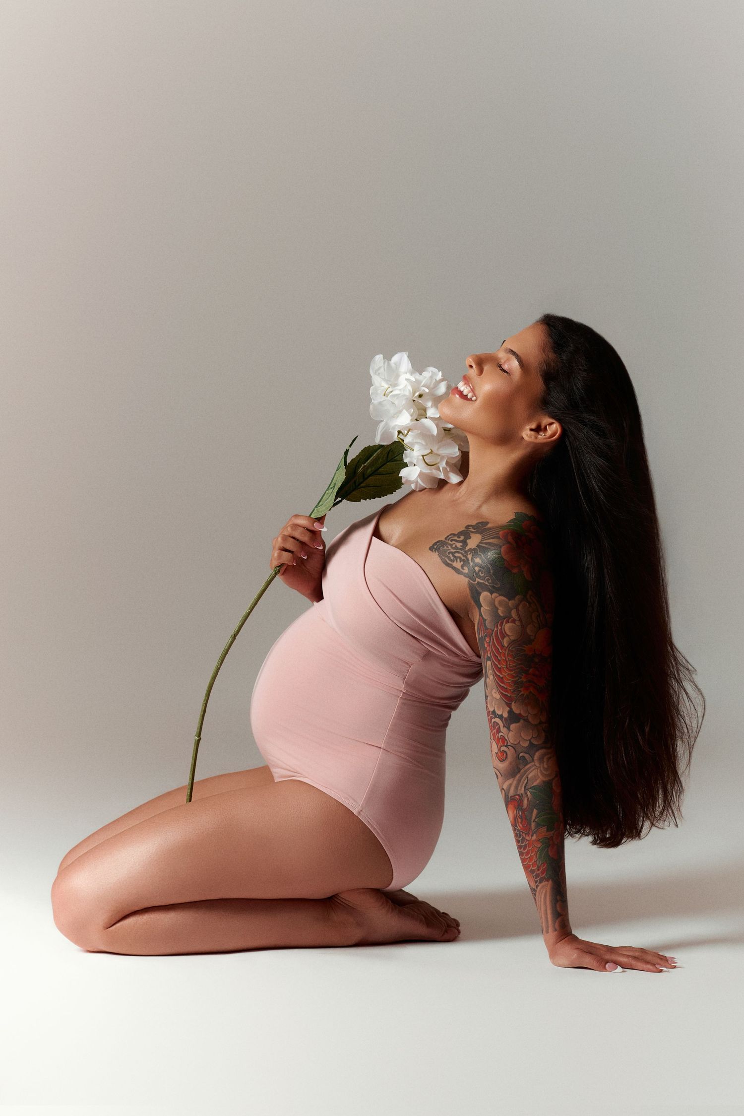 Pregnant woman with tattoos in pink bodysuit sits gracefully holding white iris flowers, smiling with long dark hair.