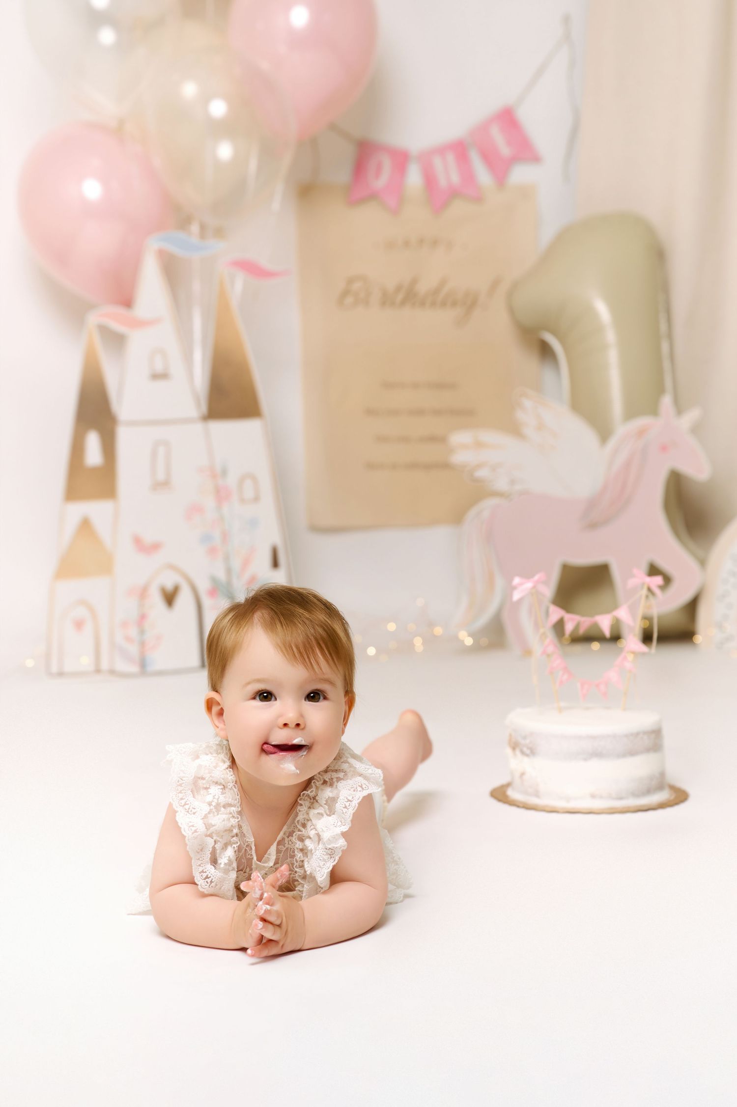 Smiling baby girl lying down at her first birthday party with pink balloons, unicorn decor, and a small white cake.