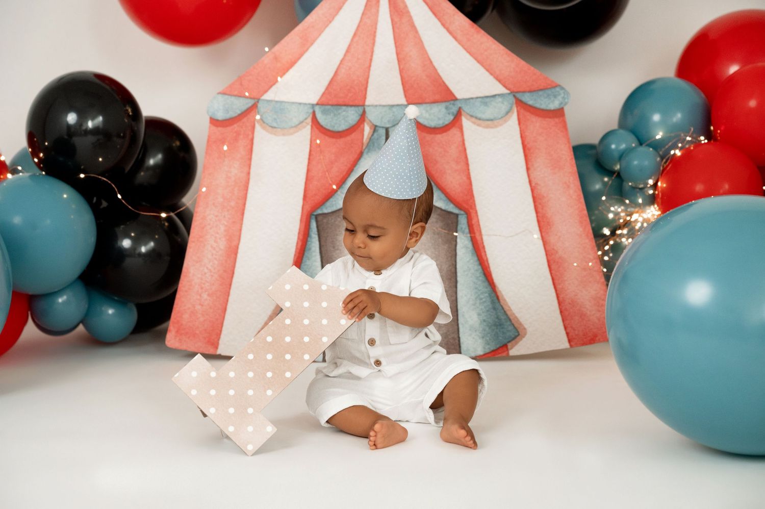 Baby boy in white outfit sits before a circus tent backdrop with red, black, and teal balloons for first birthday.