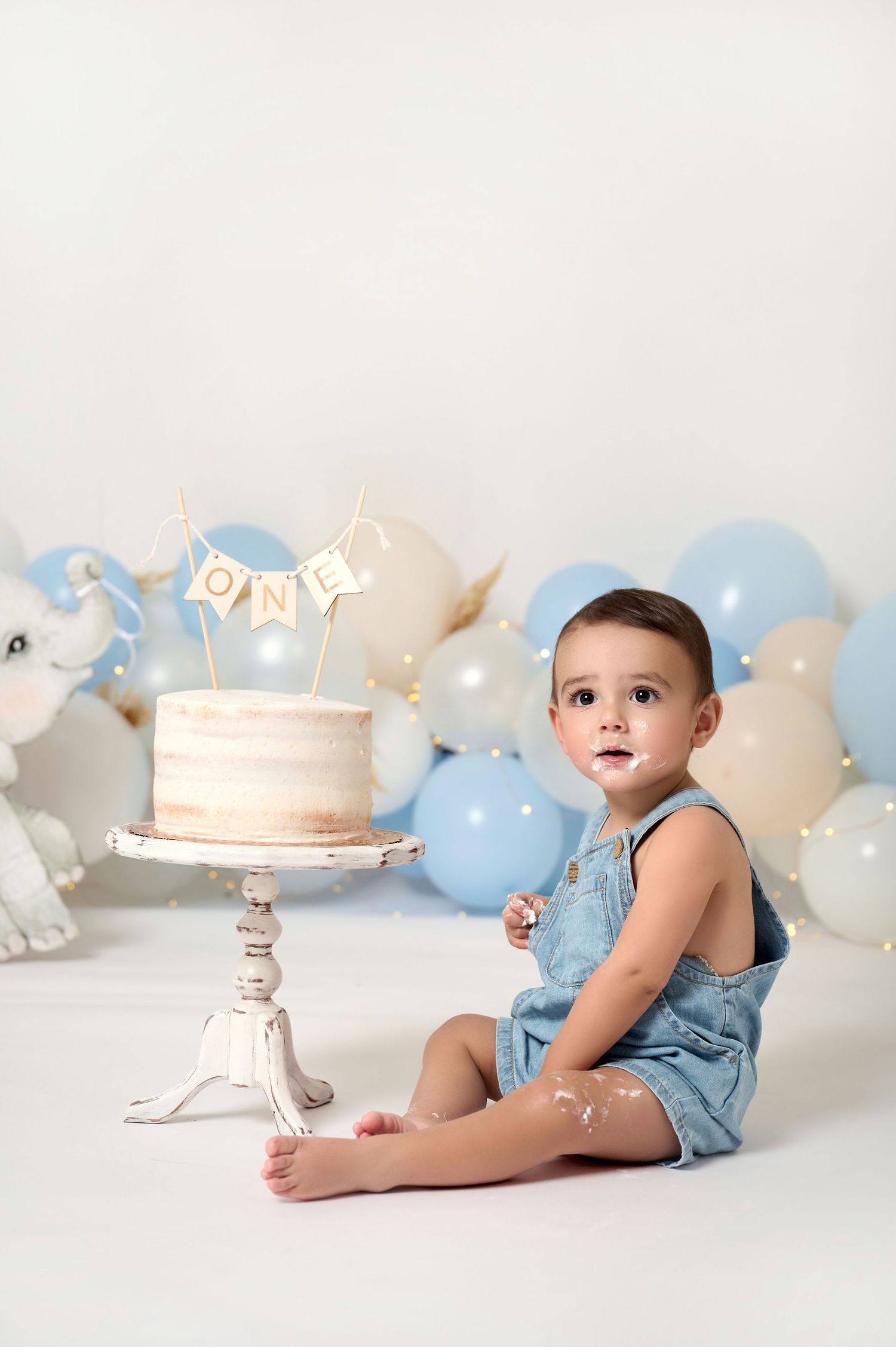 Baby boy in denim overalls sits beside a first birthday cake surrounded by blue and white balloons.