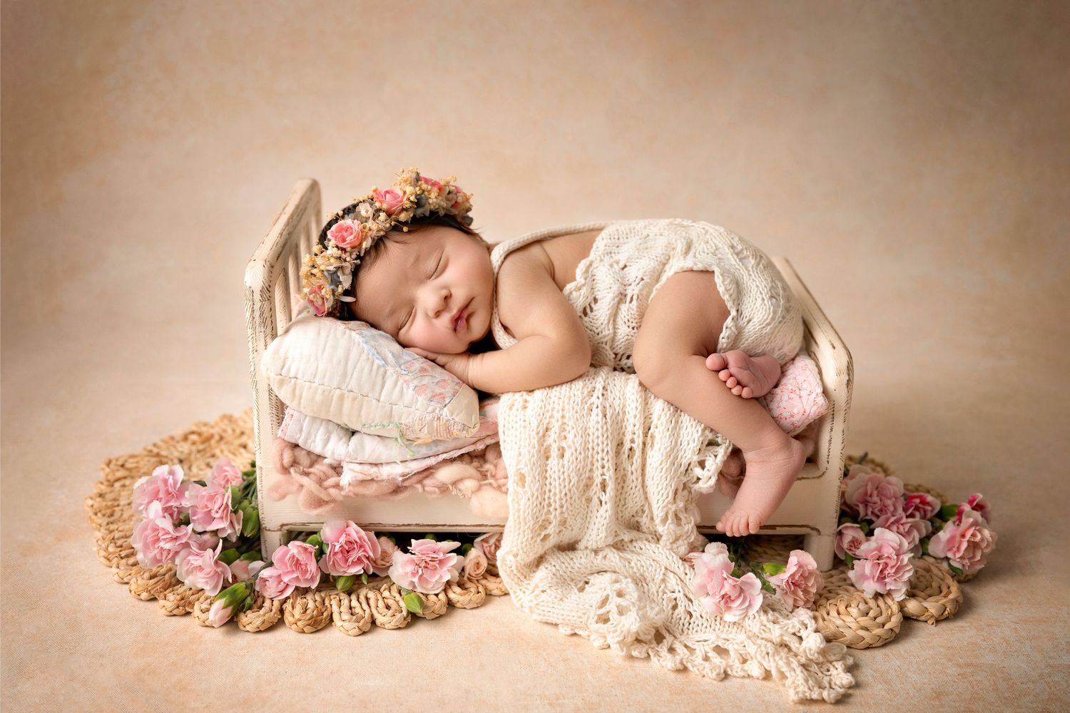 Newborn baby sleeping on a tiny vintage bed with floral wreath, lace blanket, and pink roses in a soft studio setting.