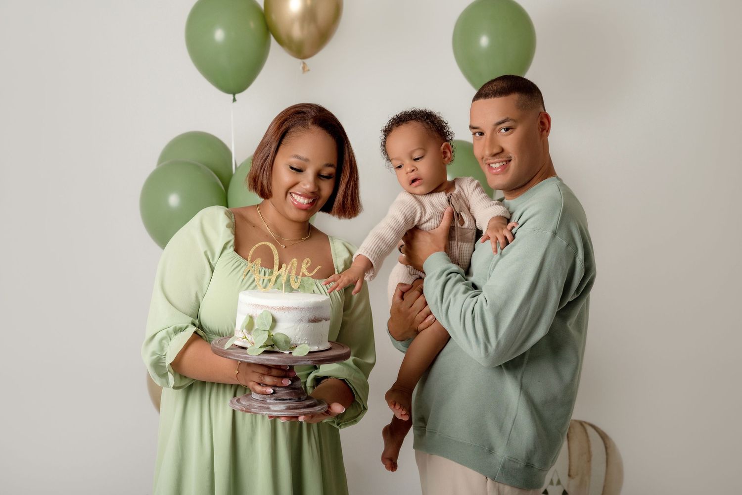 Family celebrating baby's first birthday with green balloons, mom holding cake, dad holding toddler in matching green outfits.