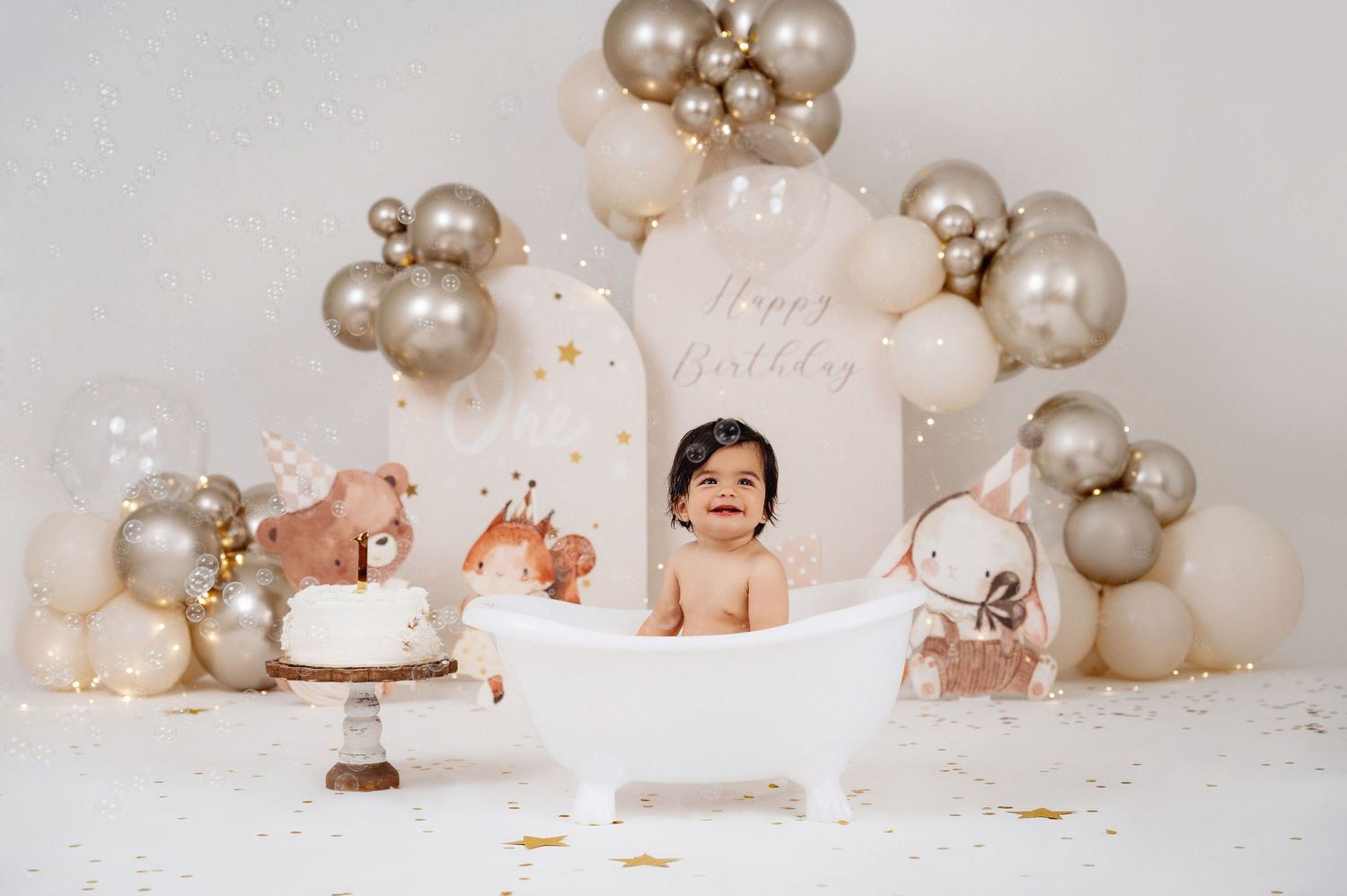 Baby sitting in a white bathtub surrounded by gold and beige balloons with a birthday cake for a first birthday cake smash.