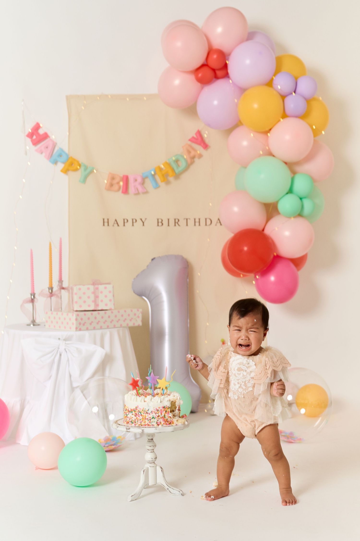 Crying baby girl celebrates first birthday with colorful balloons, cake, and festive decorations at a party photoshoot.