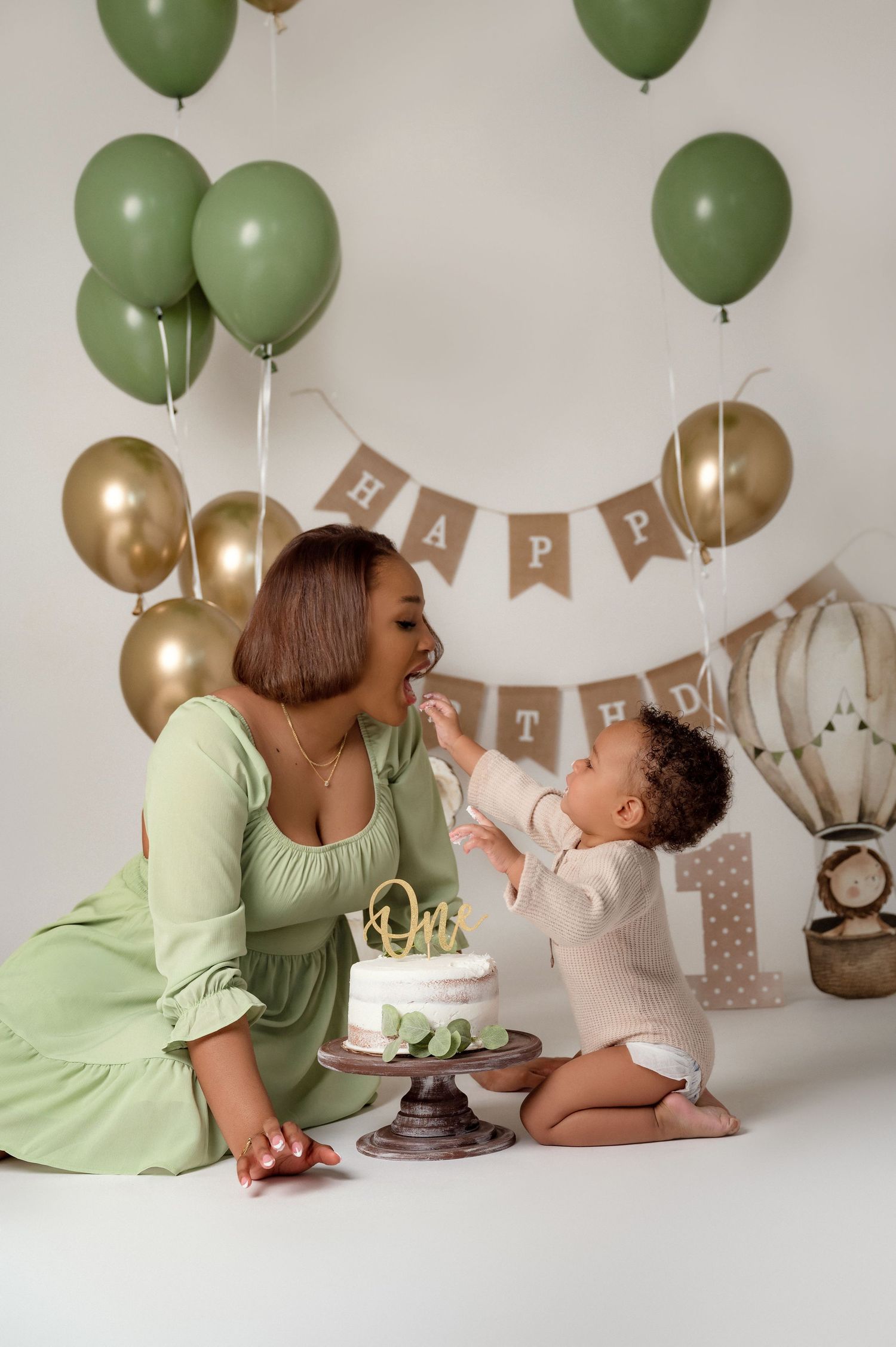 A mother in a green dress celebrates her baby's first birthday with a white cake, surrounded by green and gold balloons.