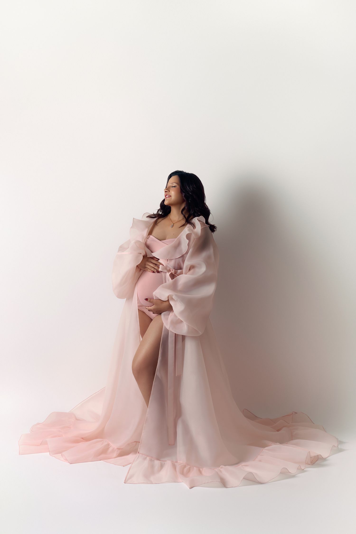 Pregnant woman in flowing pink chiffon maternity gown cradling baby bump against white studio background.