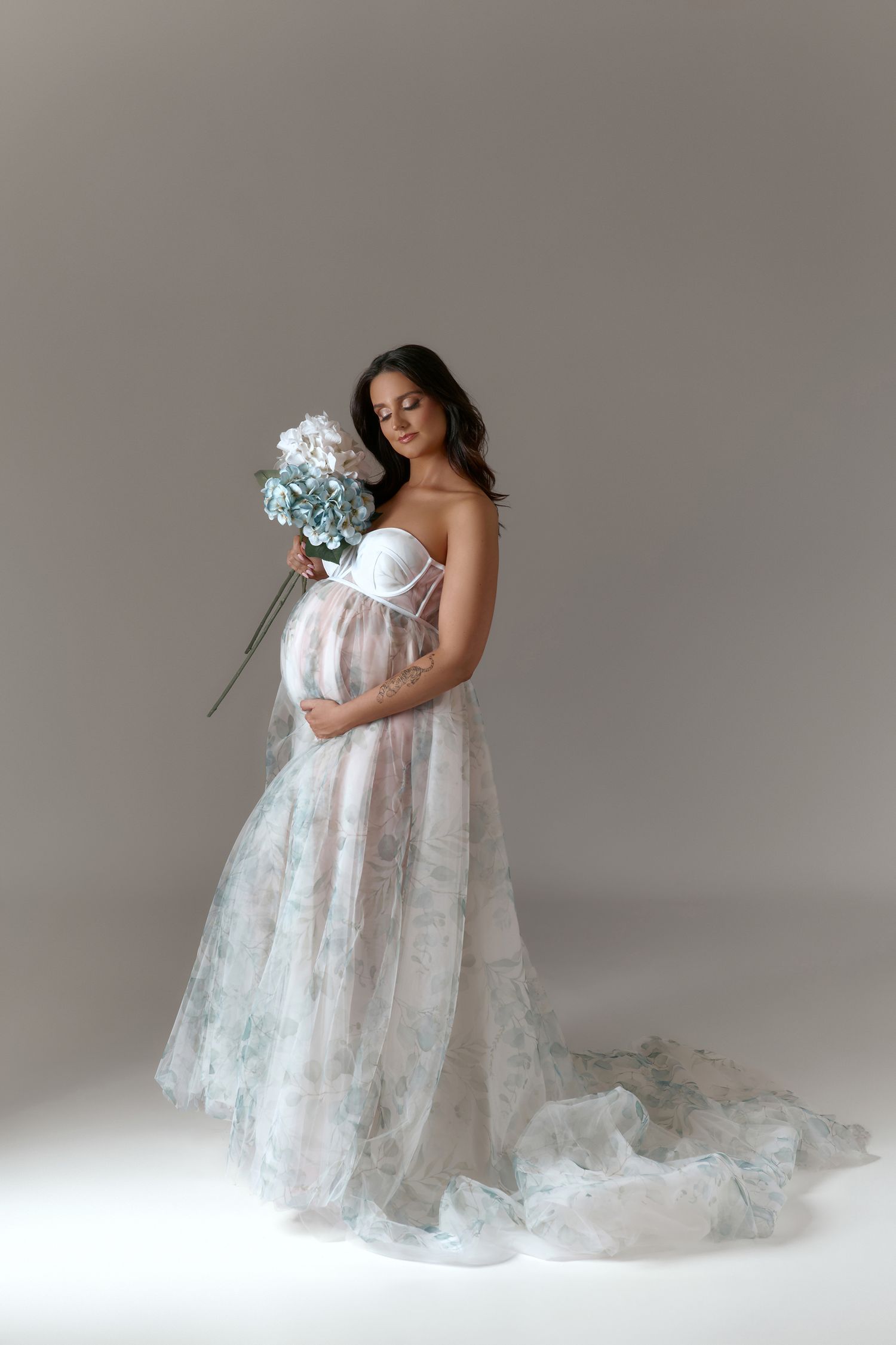 Pregnant woman in floral tulle gown holding blue and white hydrangeas, posing gracefully in a studio setting.