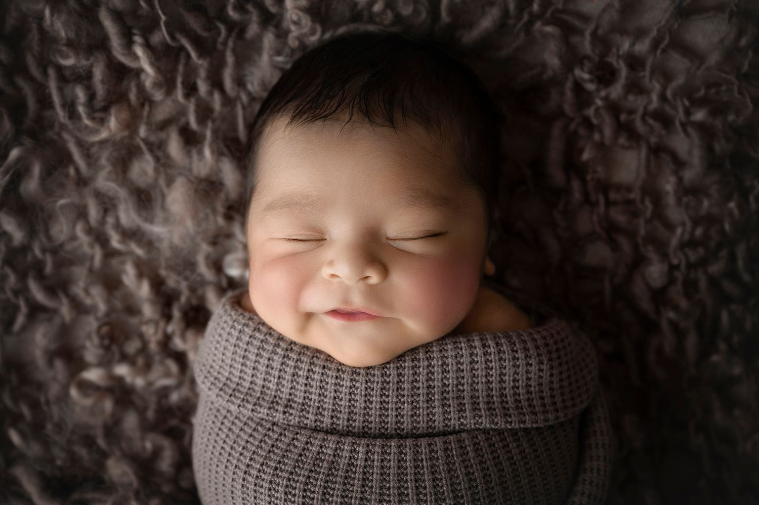 Peaceful sleeping newborn baby swaddled in gray knit wrap, smiling softly on fluffy textured blanket.