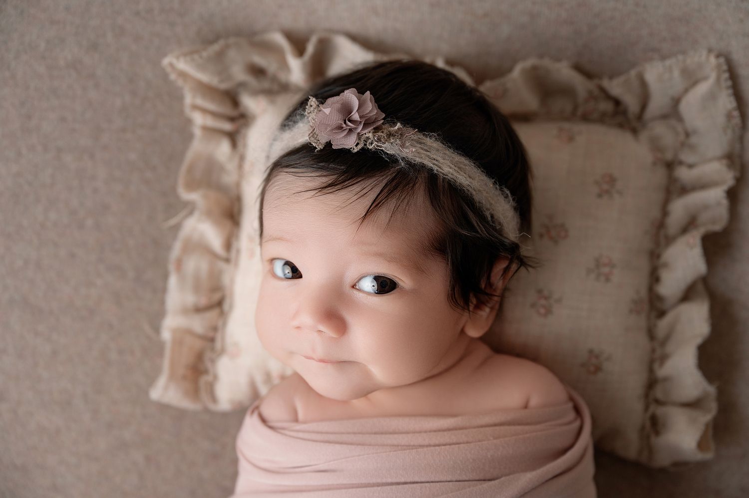Newborn baby girl wrapped in pink swaddle wearing floral headband, lying on ruffled cream pillow.