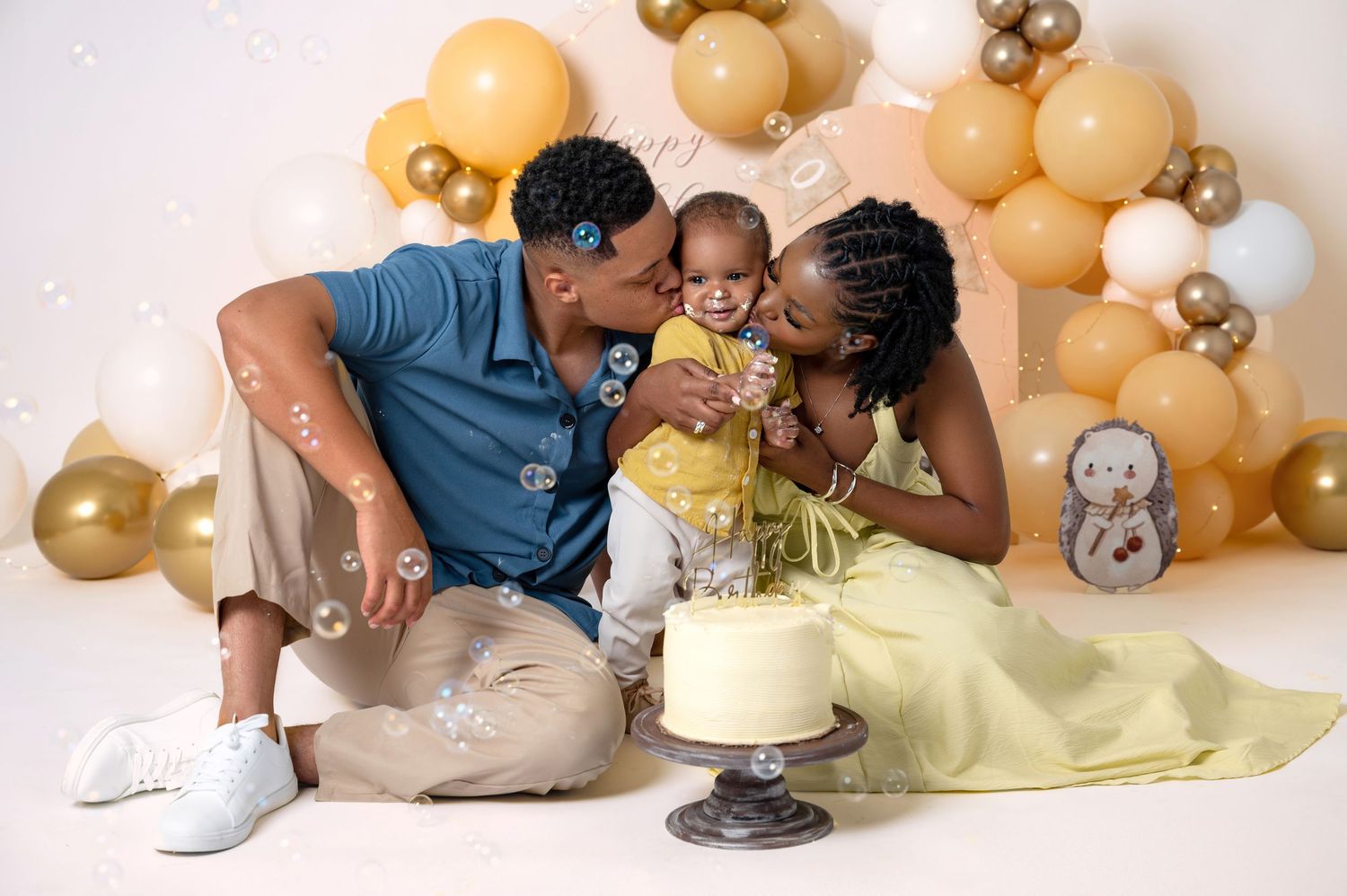 Family celebrating baby's first birthday with a white cake, gold and beige balloon arch backdrop, studio photo.