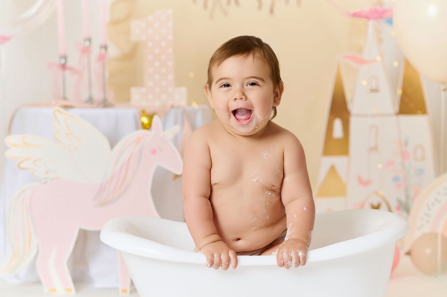 Happy baby sitting in a white mini bathtub at a unicorn-themed first birthday cake smash photo session.