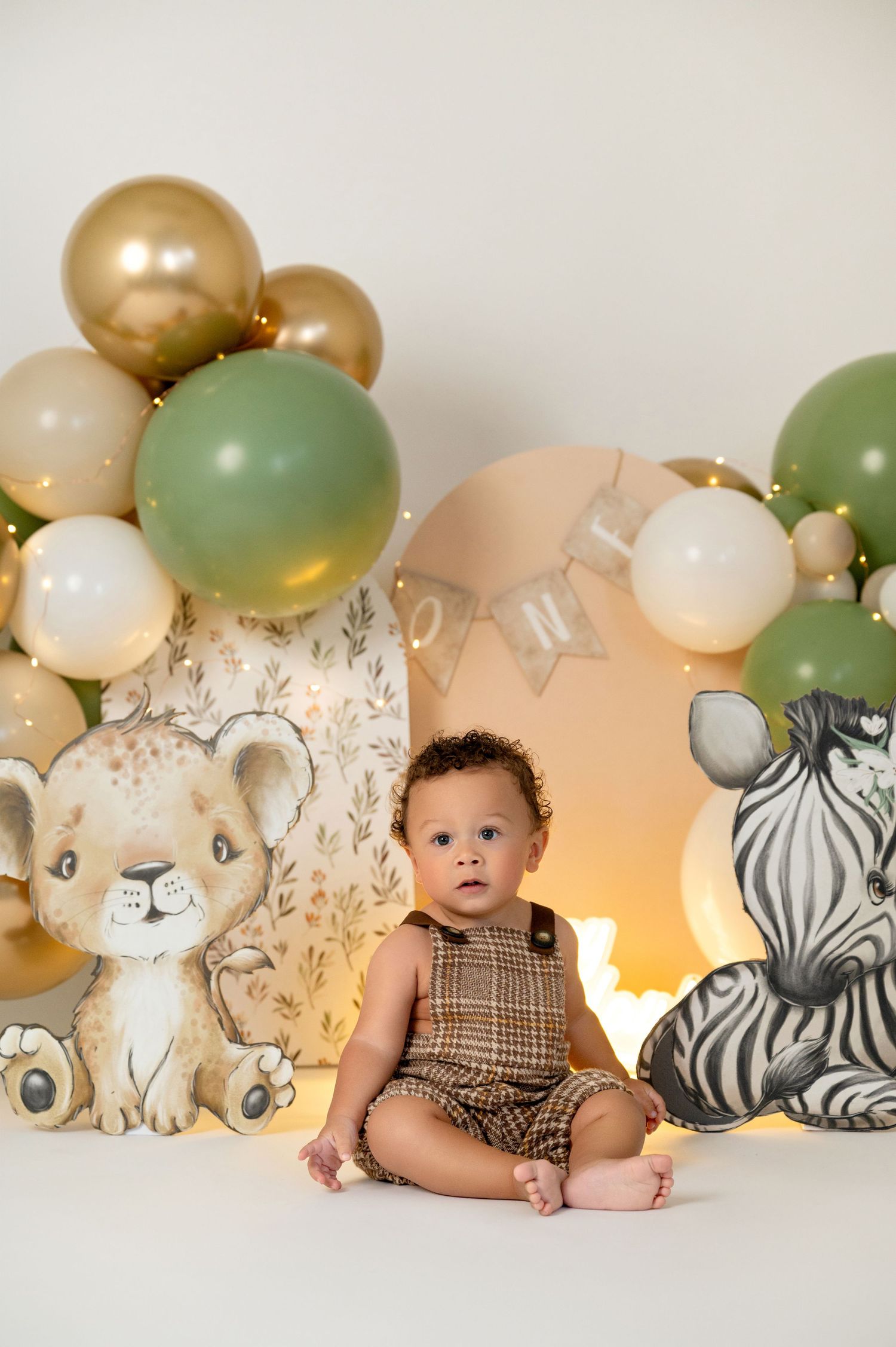 Baby boy in plaid romper sits between safari animal cutouts and green gold balloons for a jungle themed first birthday.