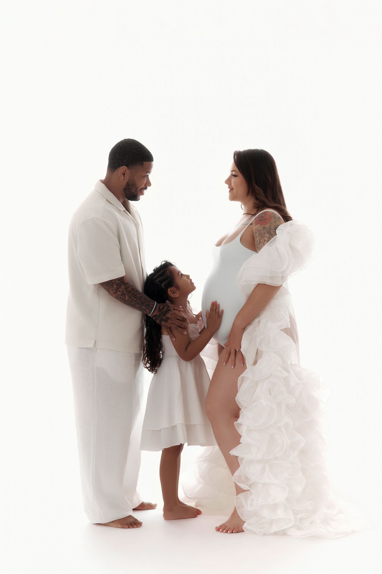 A pregnant woman in a white ruffled gown poses with her husband and daughter in matching white outfits.