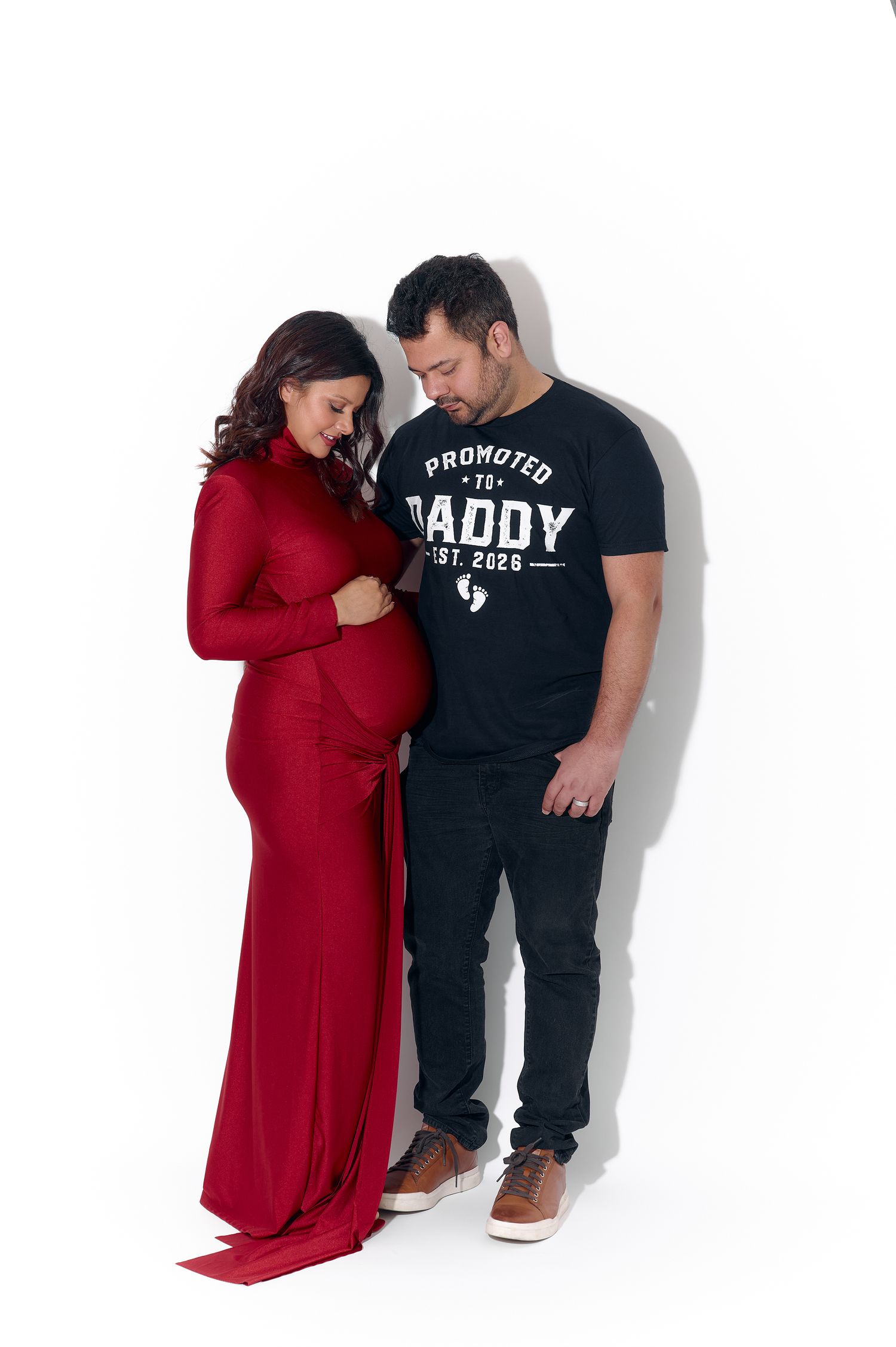 Expectant couple posing together in maternity shoot, woman in red dress, man in Daddy shirt.