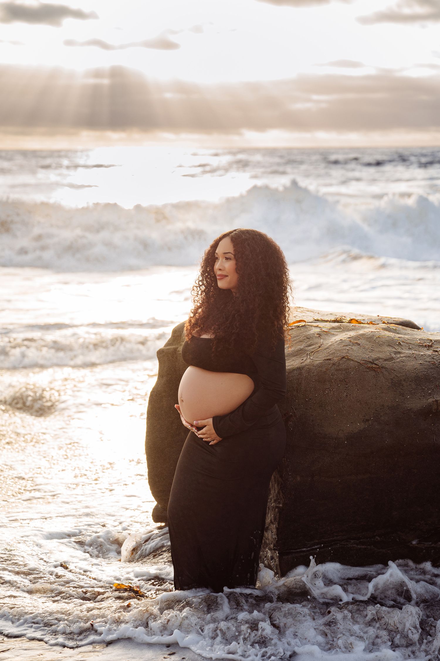 Pregnant woman in black dress cradling baby bump by ocean rocks at golden sunset.
