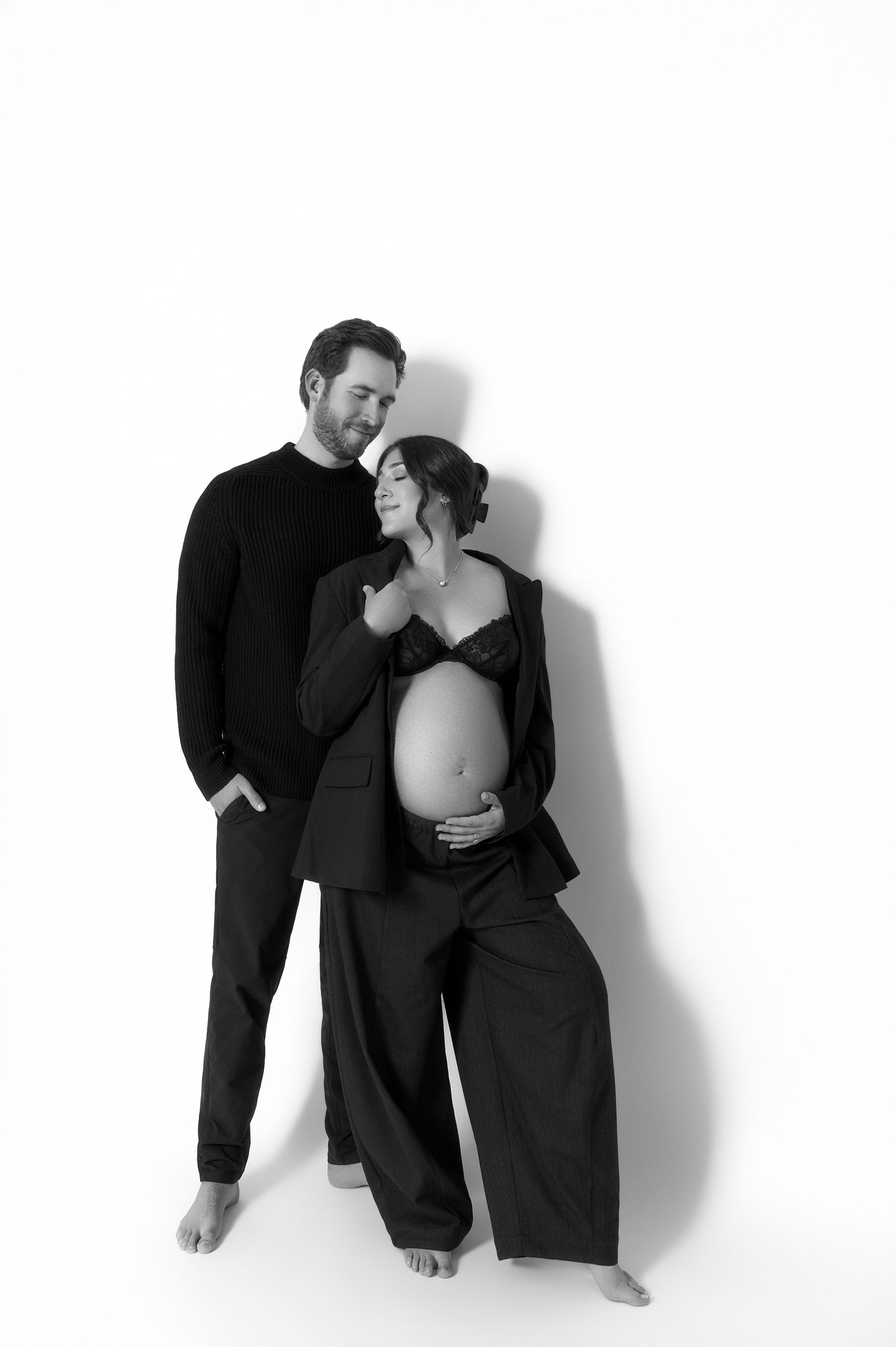 Black and white maternity photo of a couple, man embracing pregnant woman showing bare belly, studio portrait.