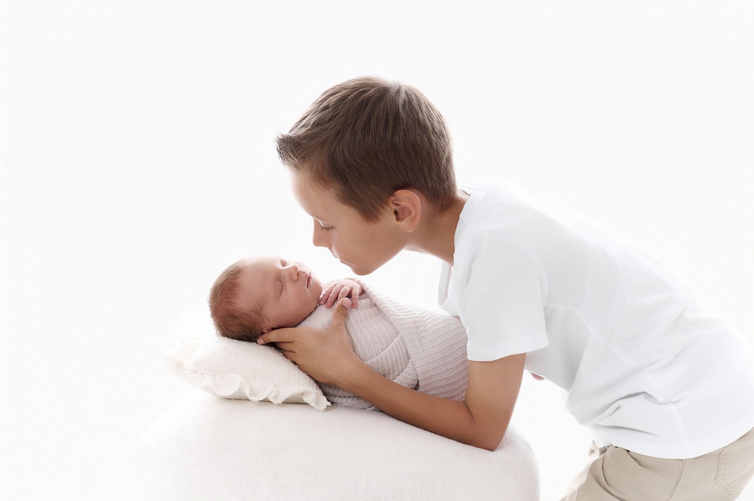 Older boy in white shirt gently kissing a swaddled newborn baby lying on a white surface.