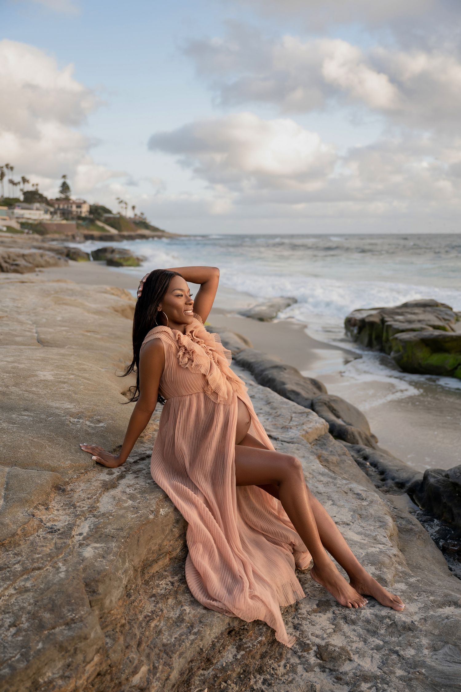 Pregnant woman in flowing peach dress posing on coastal rocks with ocean waves at sunset.