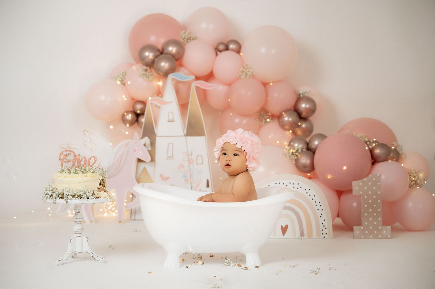Baby girl sitting in white clawfoot tub surrounded by pink and gold balloon arch for first birthday cake smash photo.