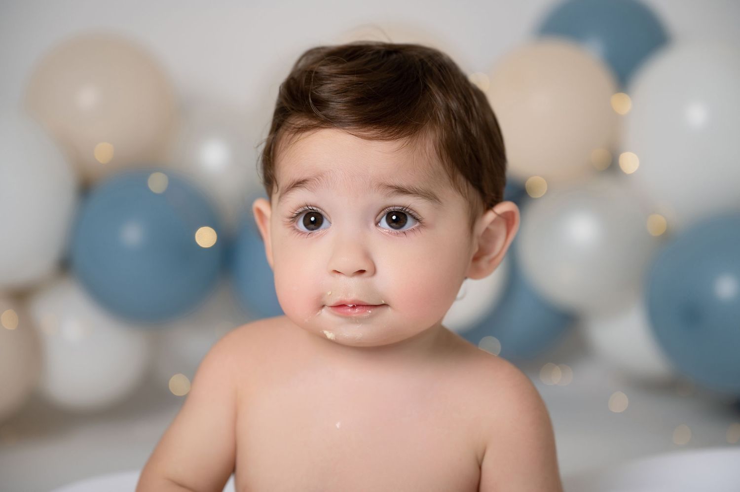 Baby boy with brown hair and big eyes sits shirtless among blue and white balloons during a first birthday cake smash.