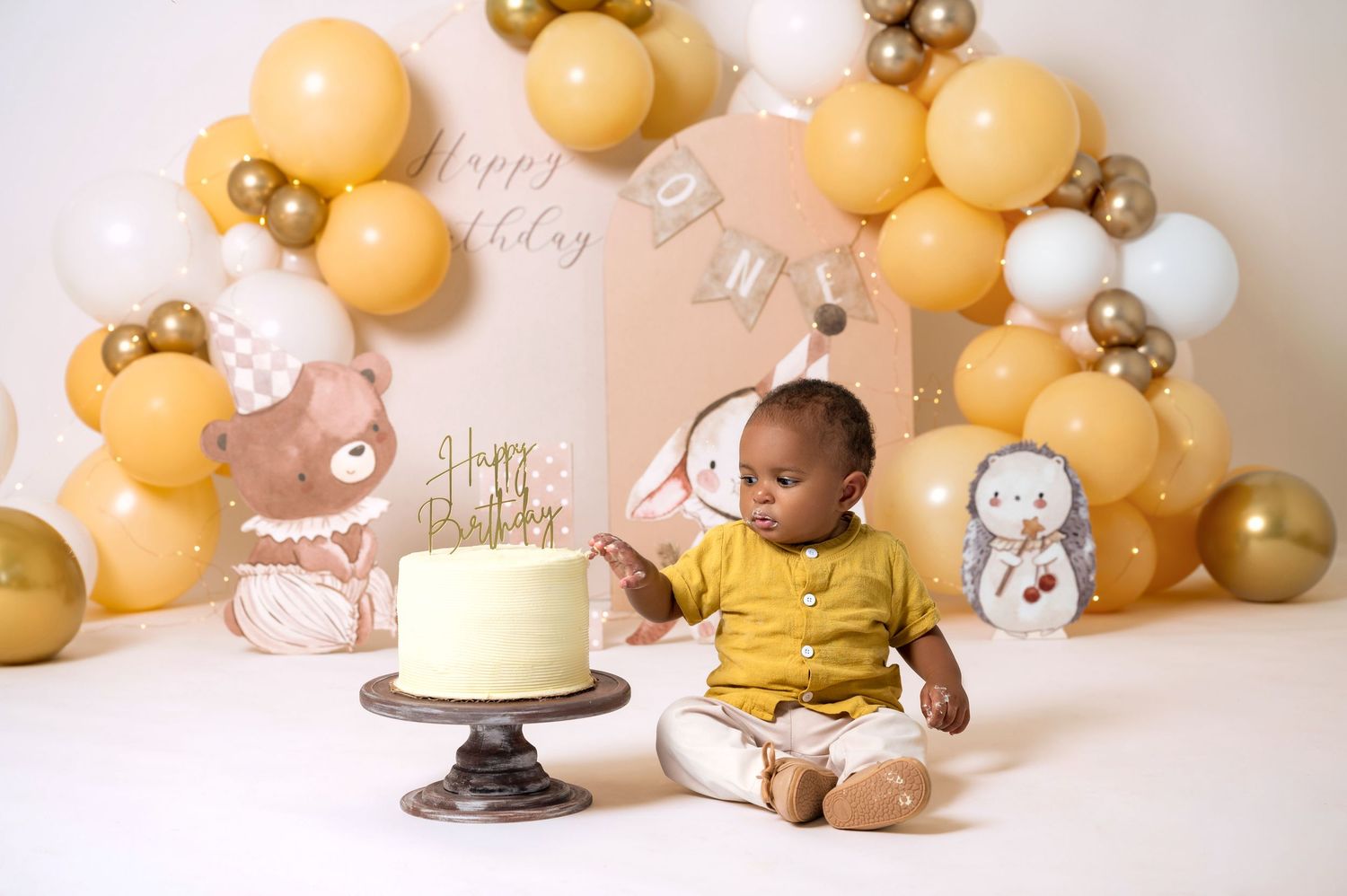 Baby's first birthday cake smash with gold and yellow balloon arch, woodland animal decorations, and white cake on wooden stand.