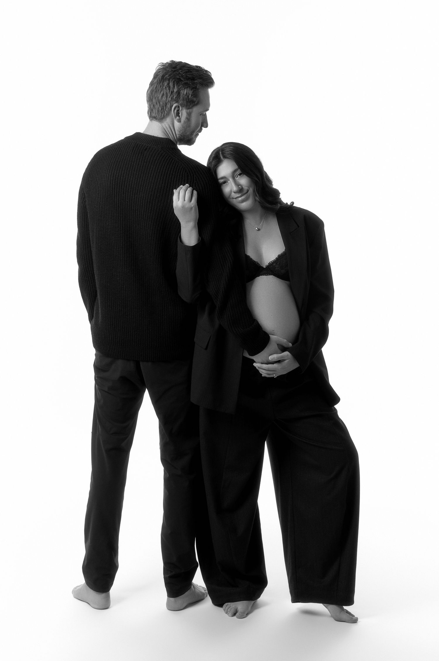 Black and white maternity photo of couple in matching black outfits embracing tenderly in studio.