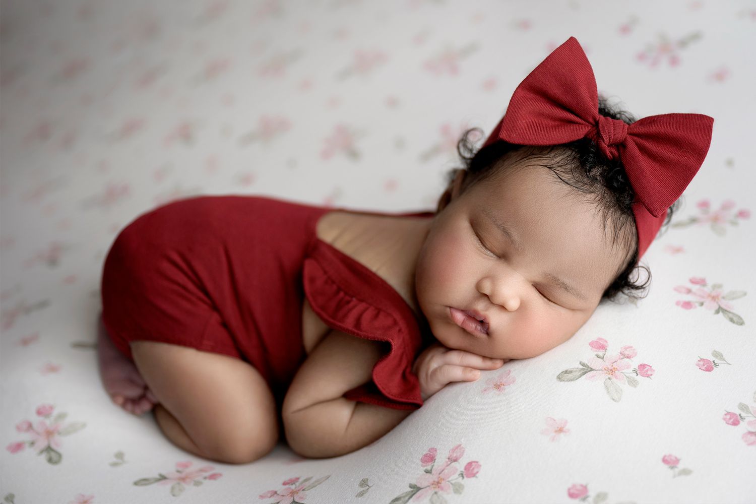 Sleeping newborn baby girl in red ruffled romper and bow headband on floral white blanket.