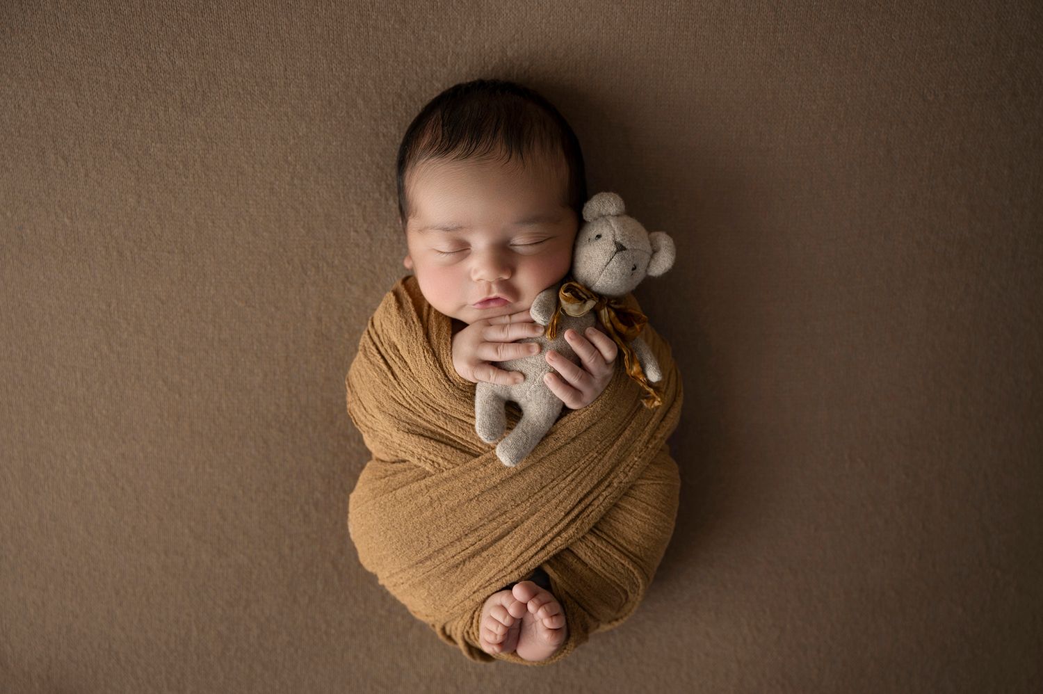 Sleeping newborn baby swaddled in golden wrap holding a tiny stuffed bear toy on brown background.