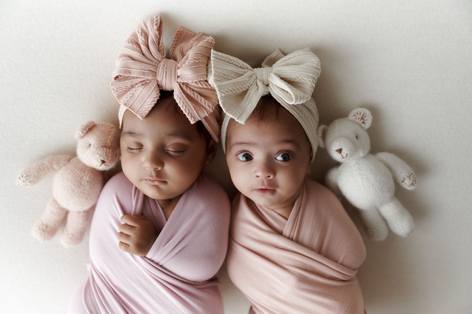 Two newborn twins wrapped in pink swaddles wear bow headbands, posed with soft teddy bears on a white background.