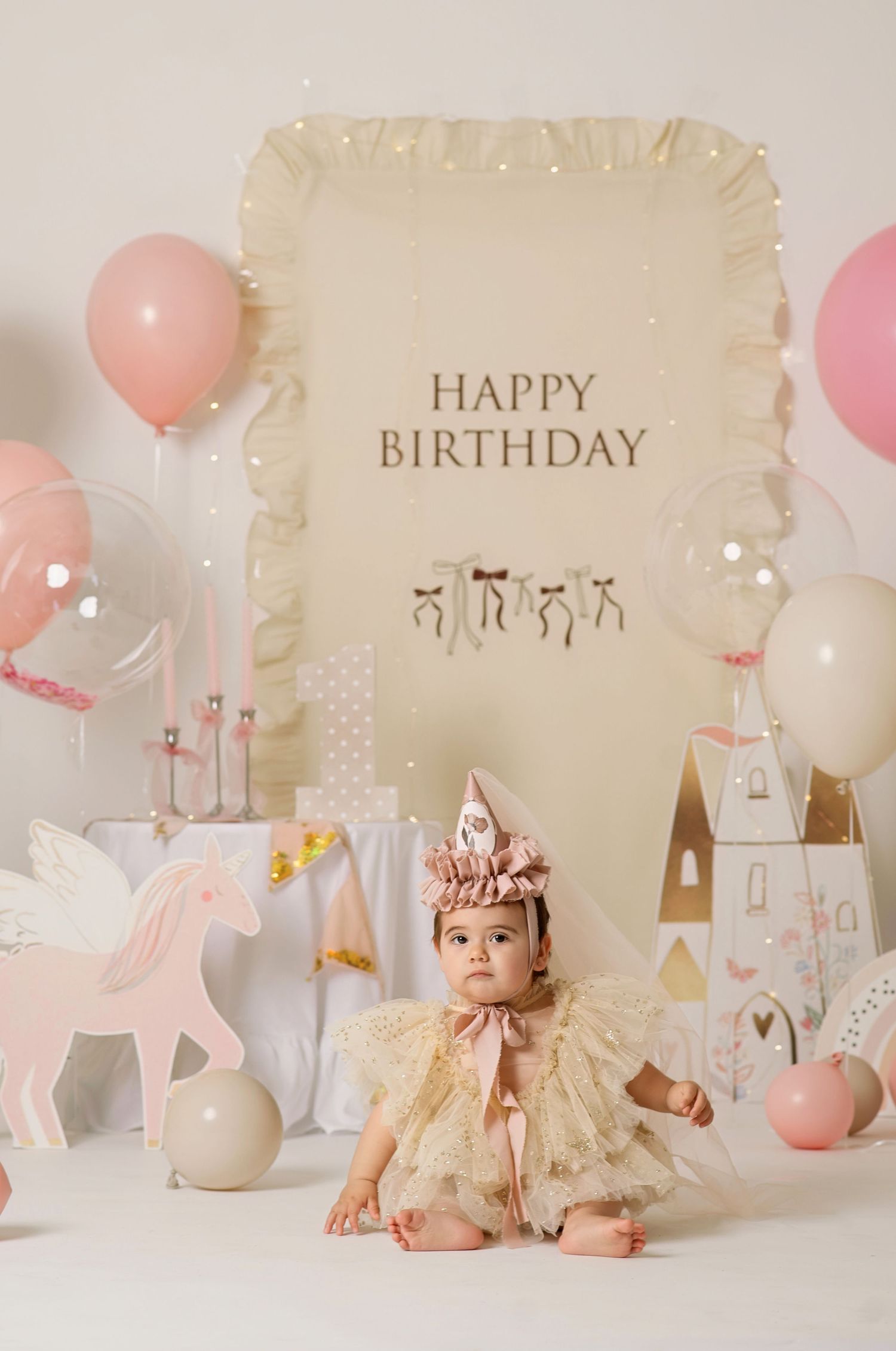 Baby girl in gold tutu dress and birthday hat sits before a Happy Birthday backdrop with pink balloons and unicorn decor.