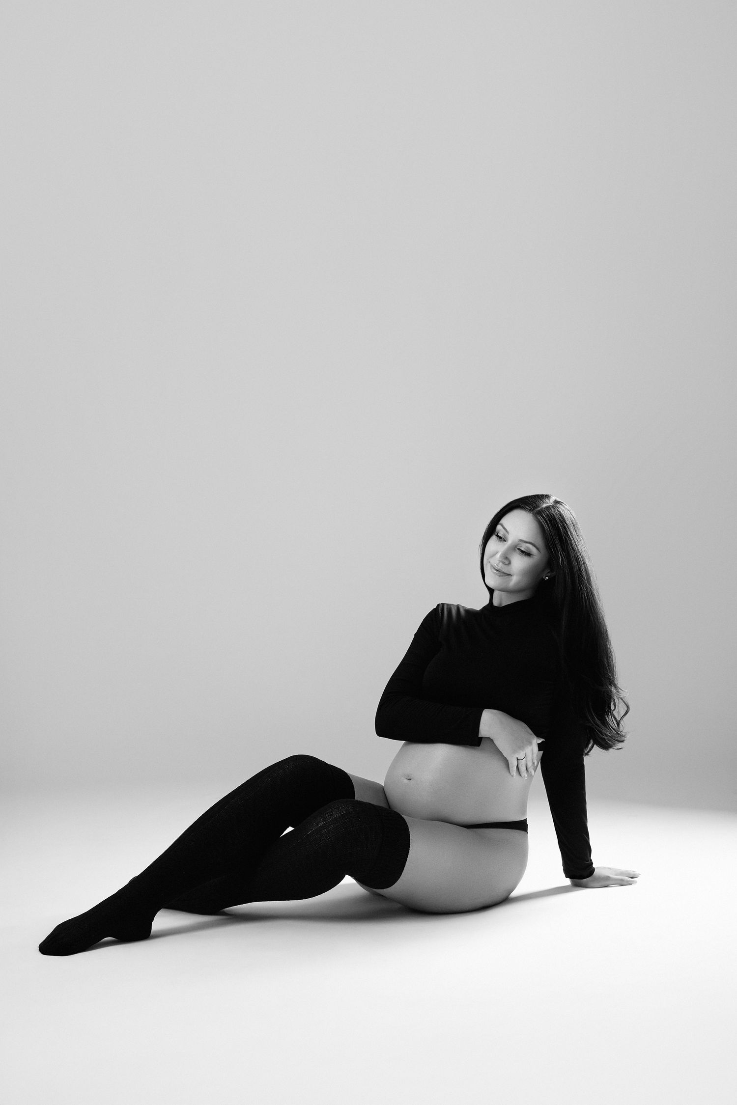 Black and white image of pregnant woman in black top and thigh-high stockings sitting on studio floor.