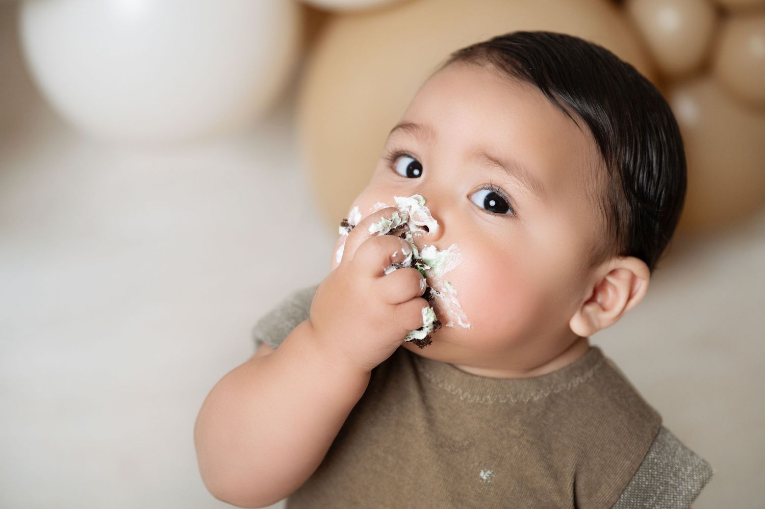 A baby boy enjoys his first birthday cake smash, covered in white frosting, with balloon decorations in the background.