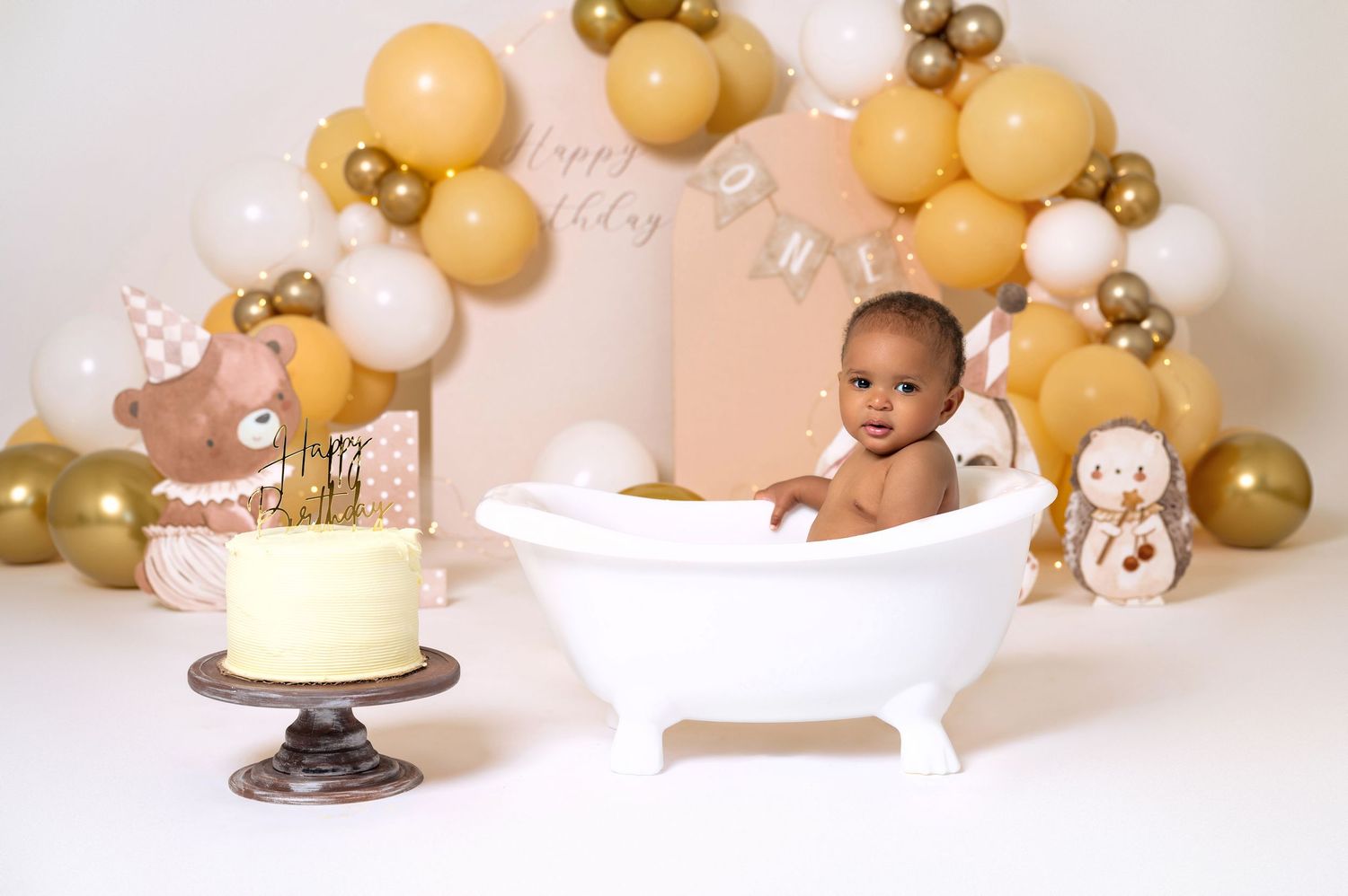 Baby sitting in white clawfoot tub with gold balloon garland and birthday cake for first birthday photo shoot.