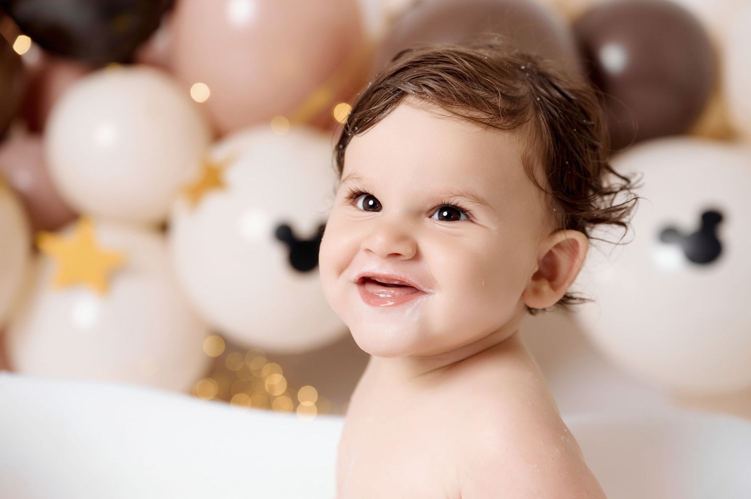 Smiling baby surrounded by Mickey Mouse balloons in pink, cream, and gold for a first birthday celebration.