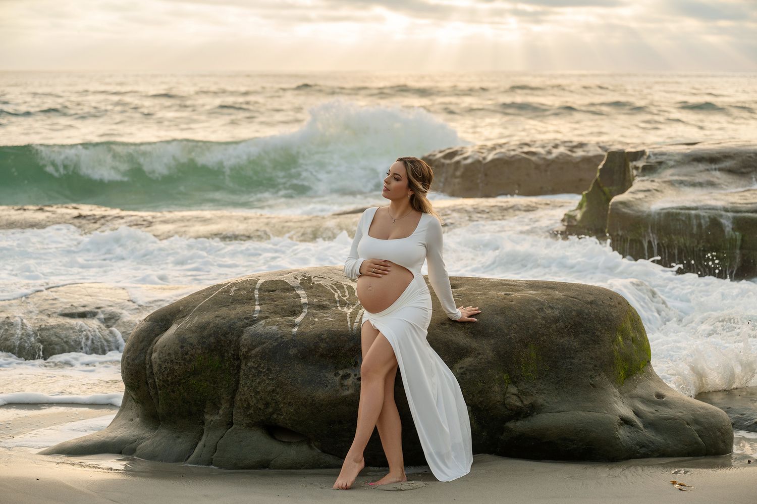 Pregnant woman in white wrap dress leans against mossy rock as ocean waves crash behind her at sunset.
