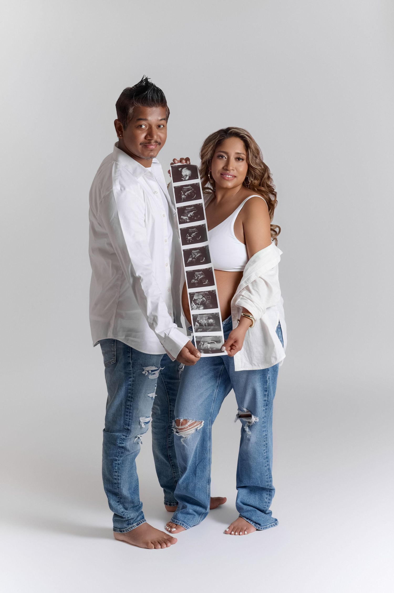 Expecting couple in jeans holding ultrasound strip, woman in white bra top showing baby bump, studio maternity photo.