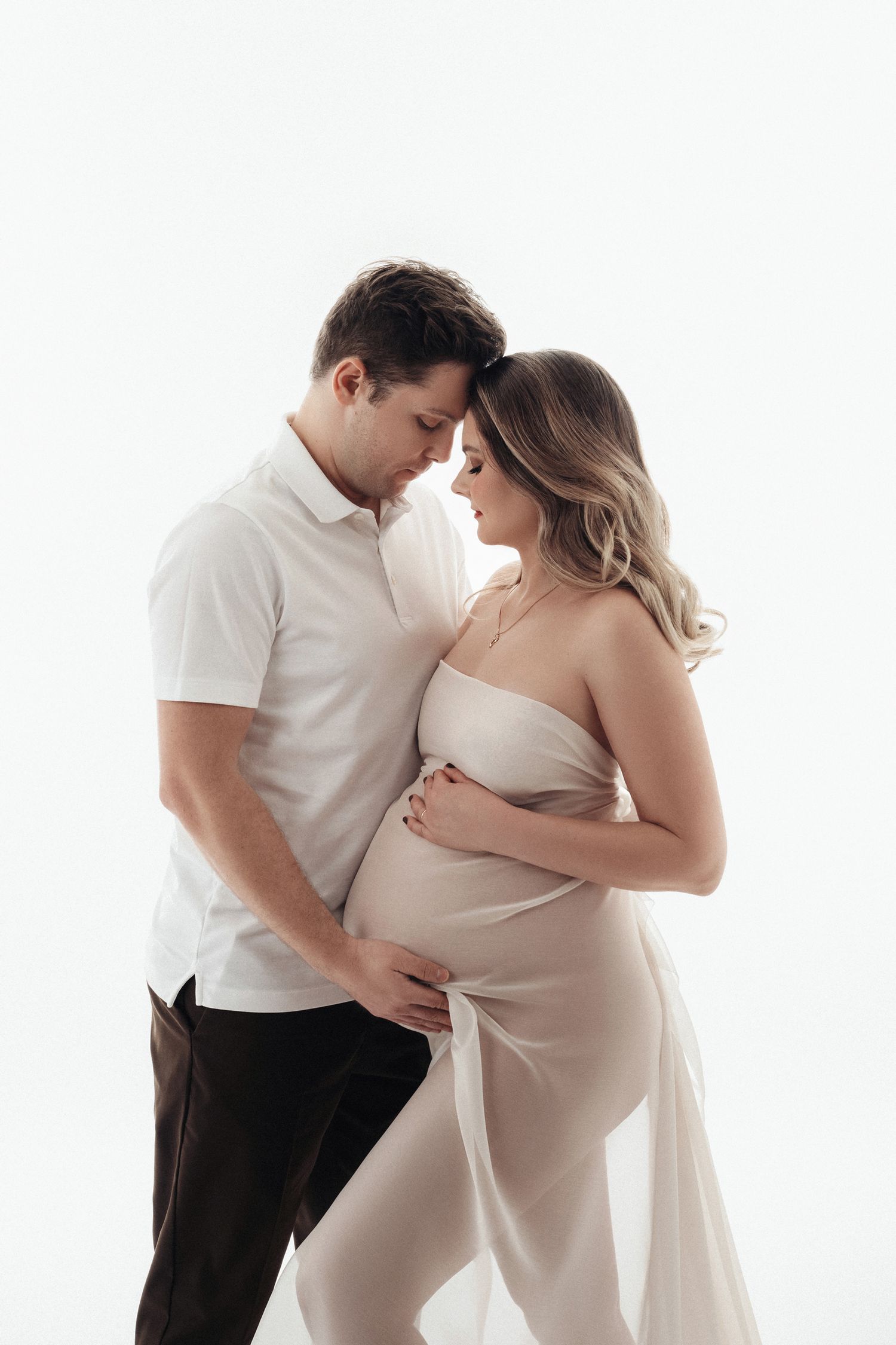 Couple sharing tender forehead touch while man cradles pregnant partners bump in white studio photo.