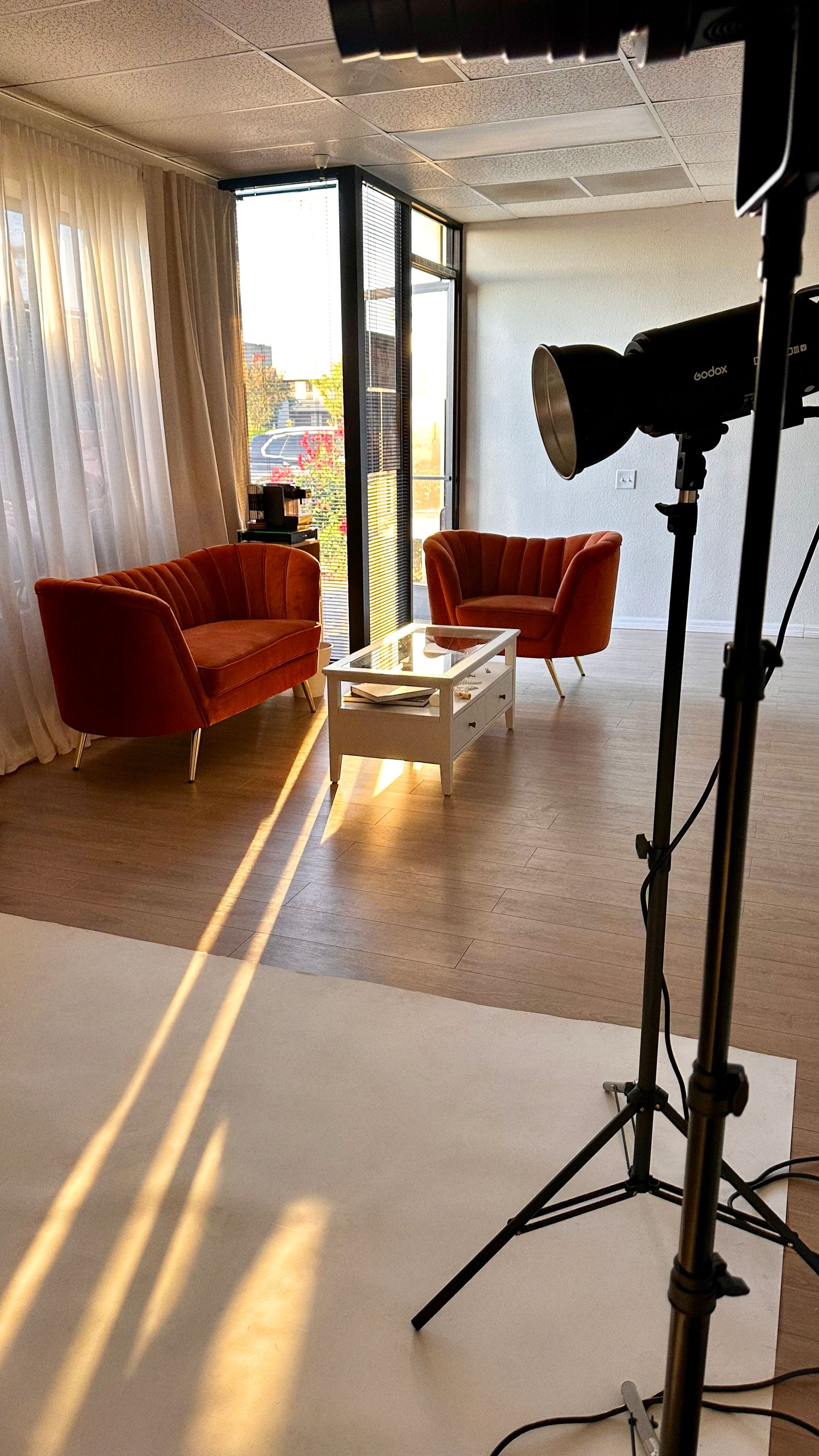 Photography studio setup with orange velvet chairs, white coffee table, hardwood floors, and professional studio lighting on tripod.