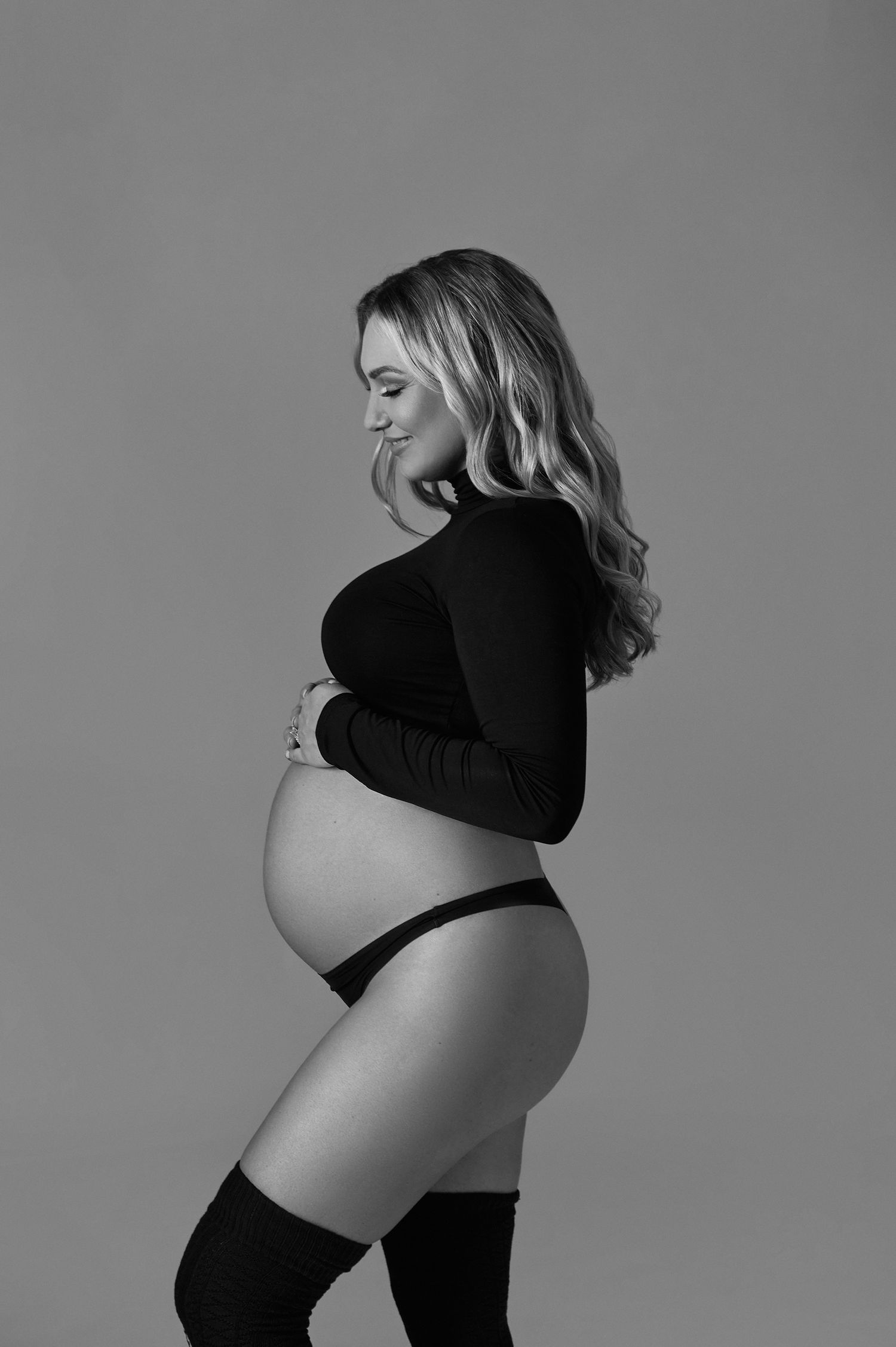 Black and white maternity photo of a pregnant woman in a black crop top and thigh-high socks cradling her baby bump.