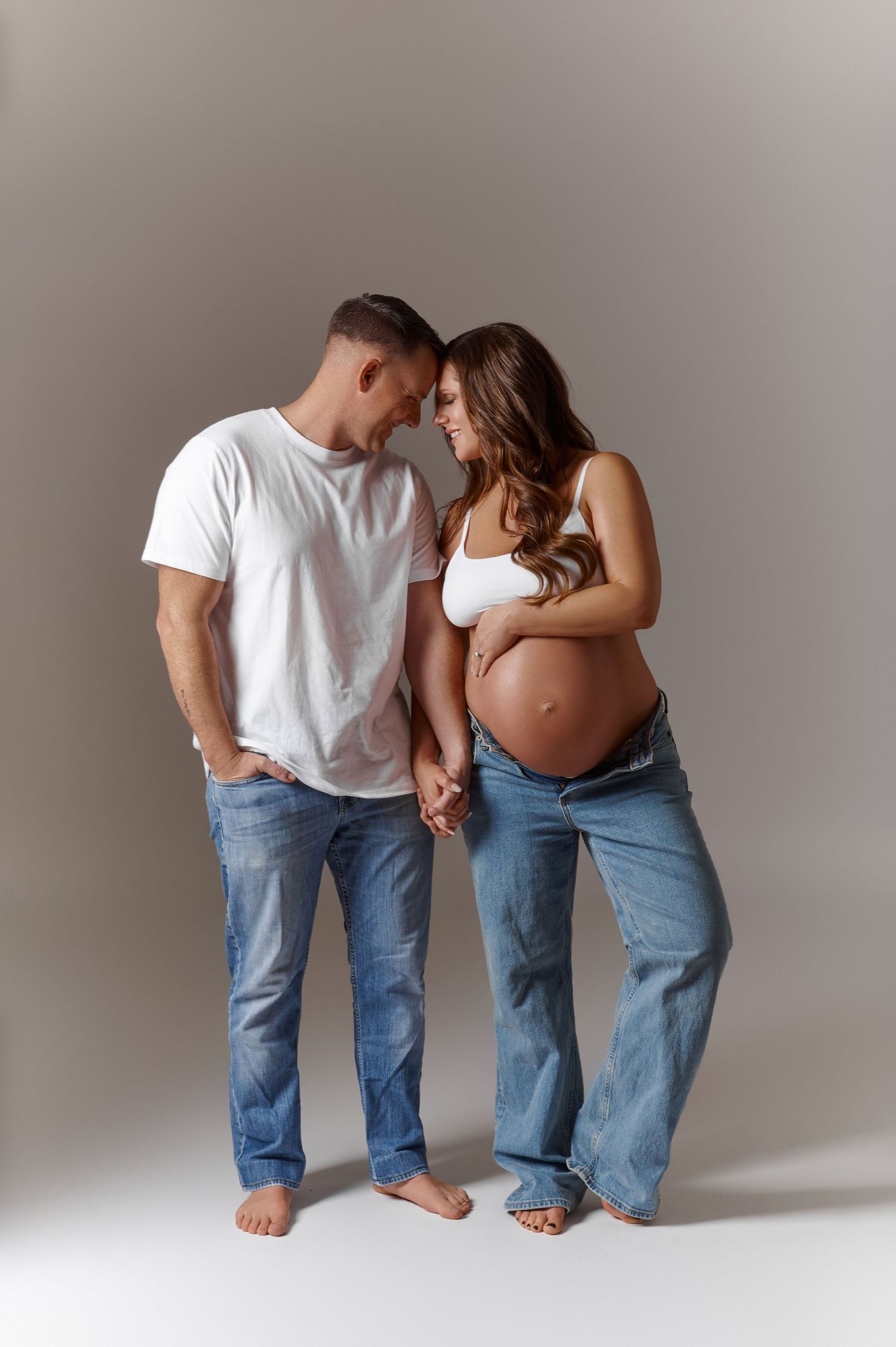 Pregnant couple holding hands in studio, man in white tee and jeans, woman in white crop top showing baby bump.