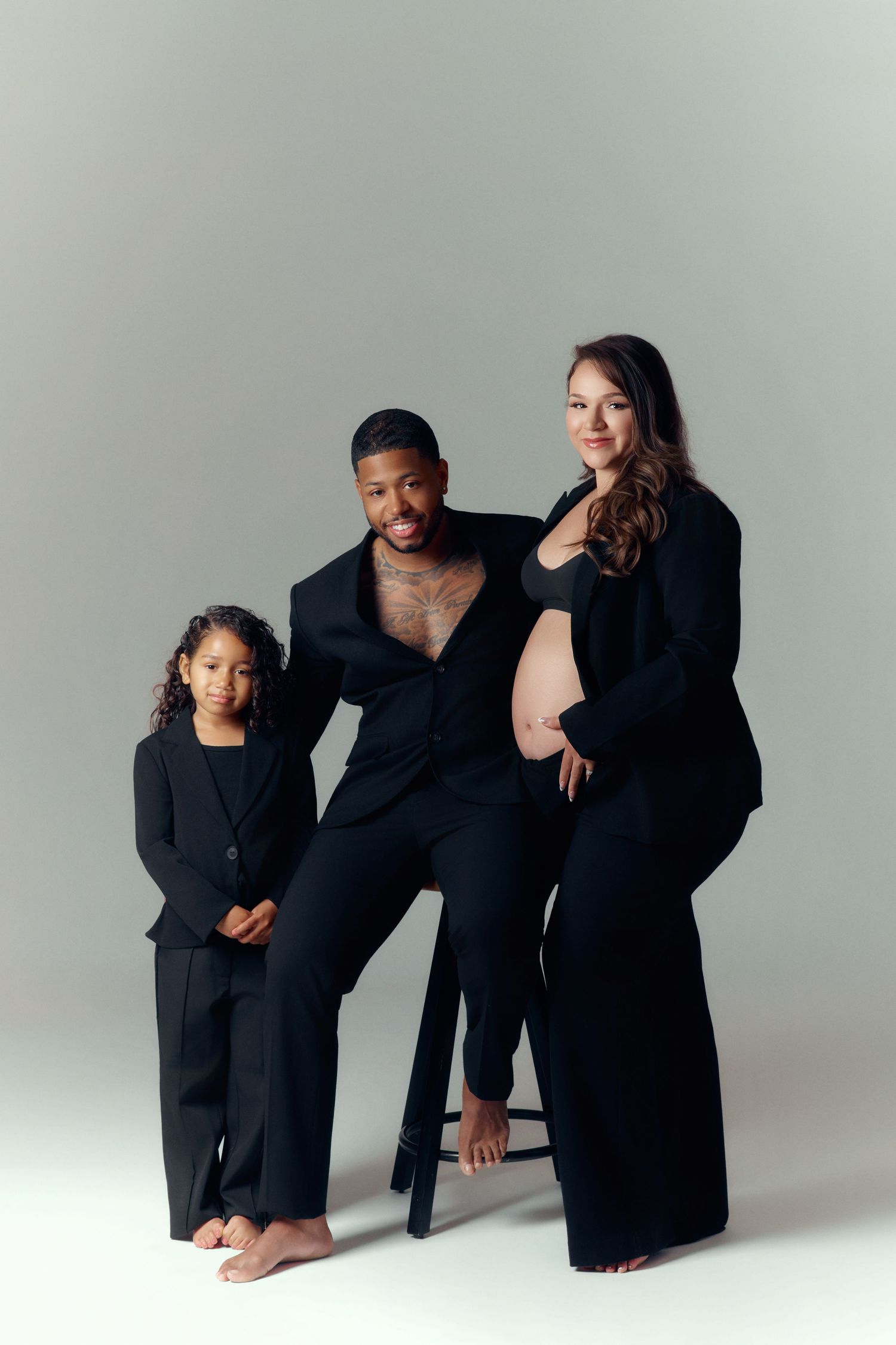 A family maternity photo with a pregnant woman, man in black suit, and young girl, all dressed in black against a gray background.