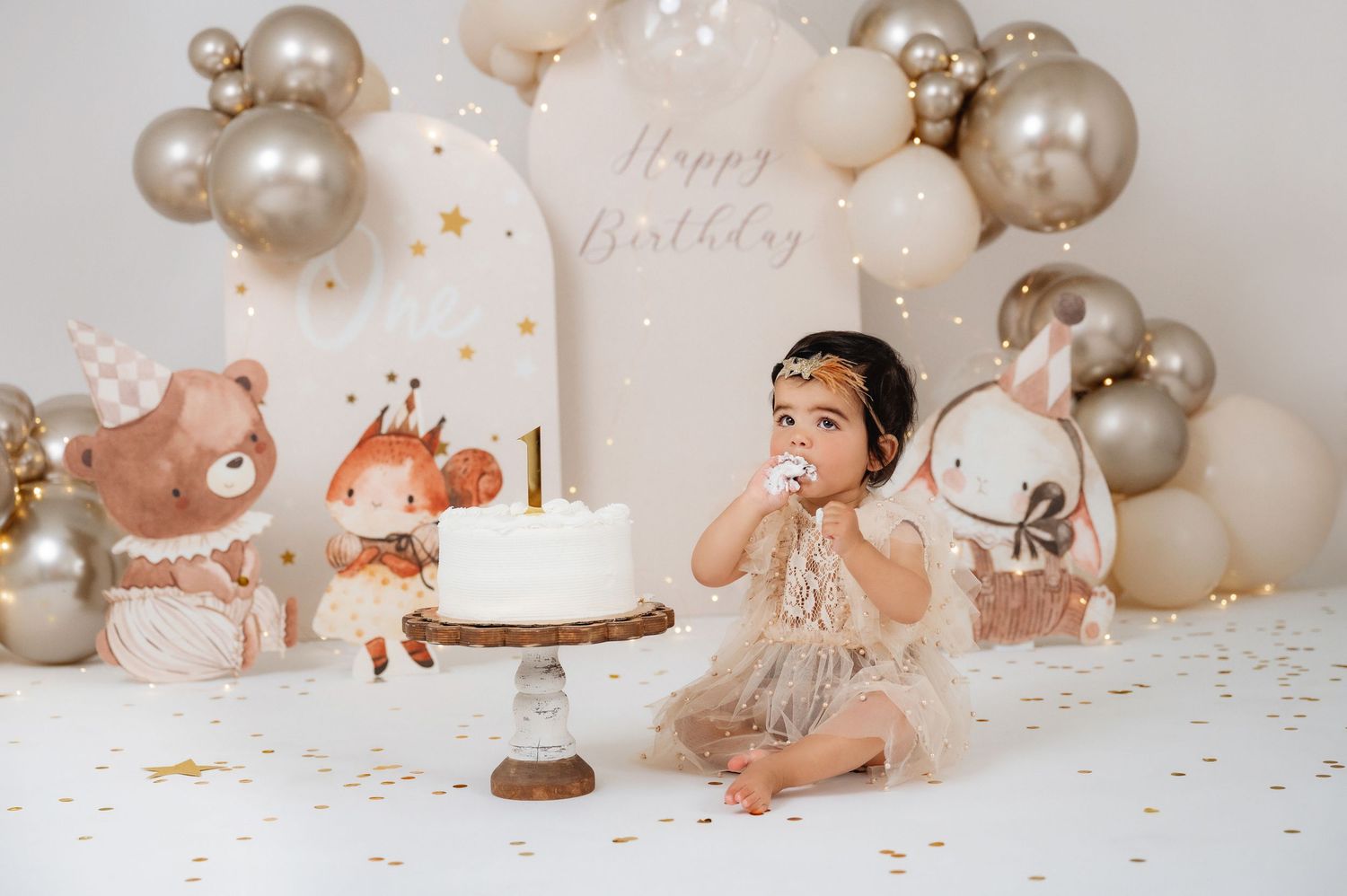 Baby girl in tutu celebrates 1st birthday smashing cake surrounded by balloon arch and woodland animal cutouts.