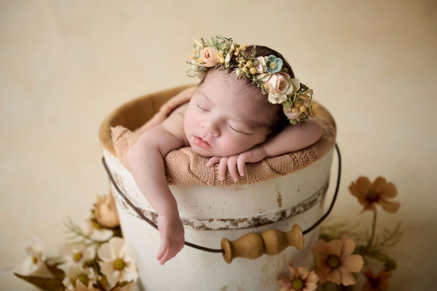 Sleeping newborn baby wearing a floral crown resting in a rustic white bucket surrounded by flowers.