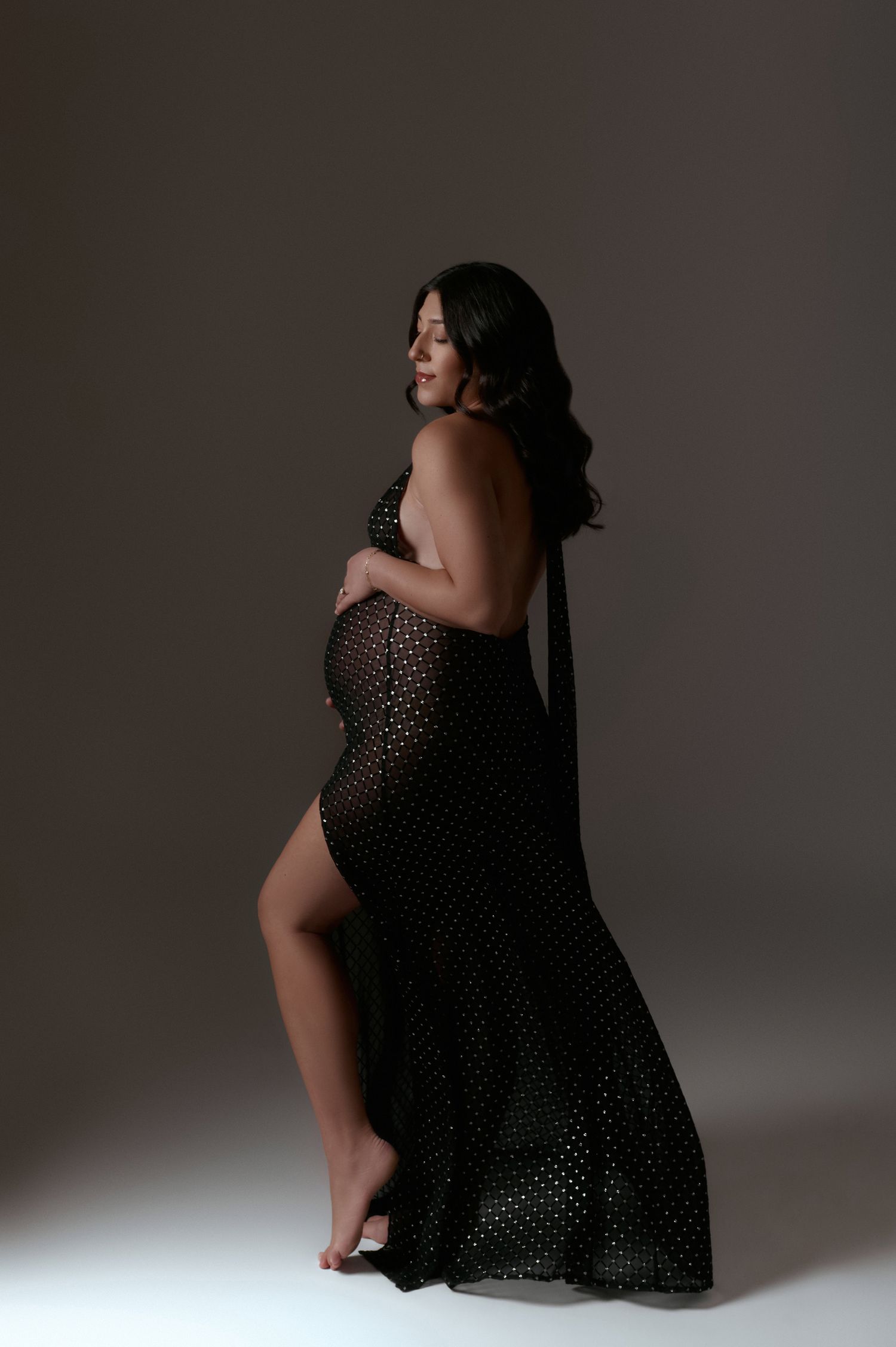 Pregnant woman in sheer black embellished gown with side slit posing in elegant maternity portrait.
